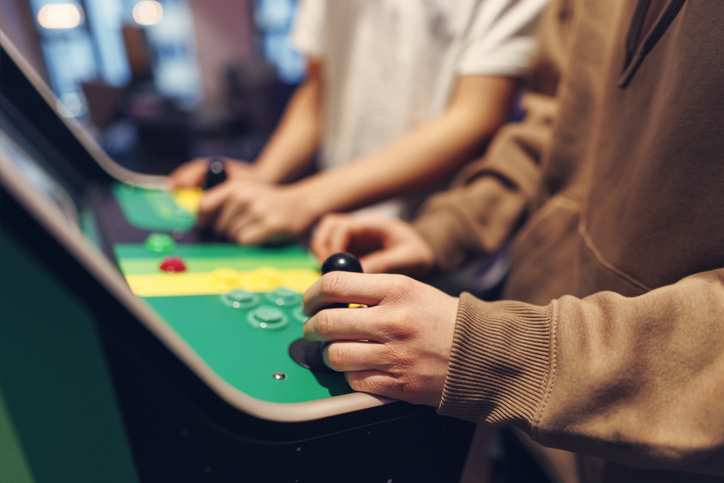 Two people, partially visible, playing a retro arcade game. One wears a casual hoodie; the other is in a t-shirt. Their hands are on the joystick and buttons