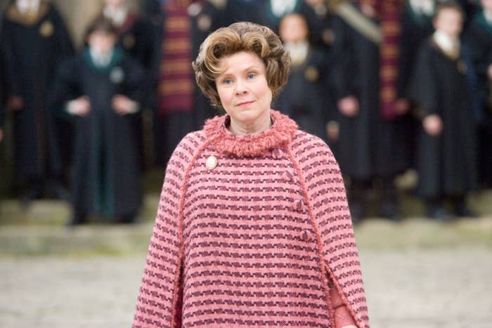 Imelda Staunton sebagai Dolores Umbridge dalam adegan Harry Potter berdiri di luar ruangan di depan orang lain dengan jubah hitam.  Dia memakai ponco bermotif dengan bros