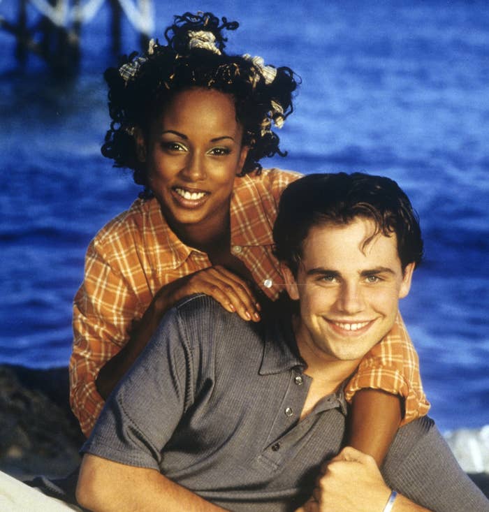 Trina McGee leans on Rider Strong's shoulder on a beach. Trina wears a checkered shirt, and Rider wears a polo and light pants