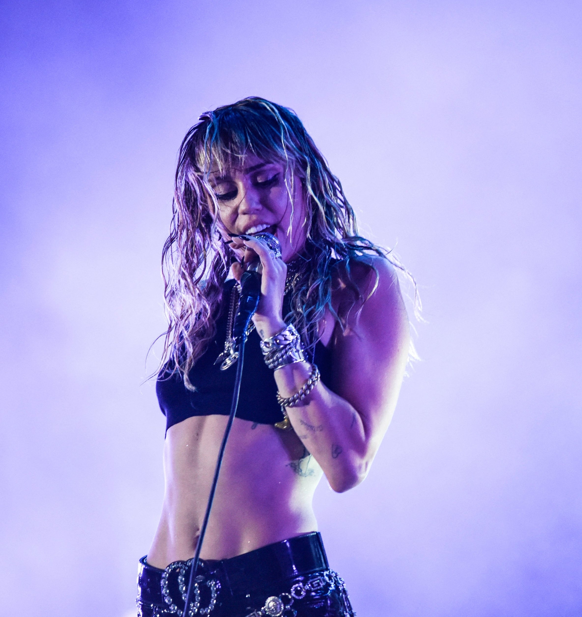 Miley Cyrus performs on stage, wearing a cropped top and adorned with layered necklaces and bracelets