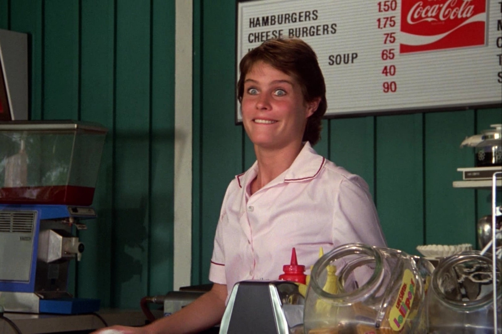 A person in a diner uniform with a name tag stands behind a counter with a joyful expression. A menu board listing food items is partially visible in the background