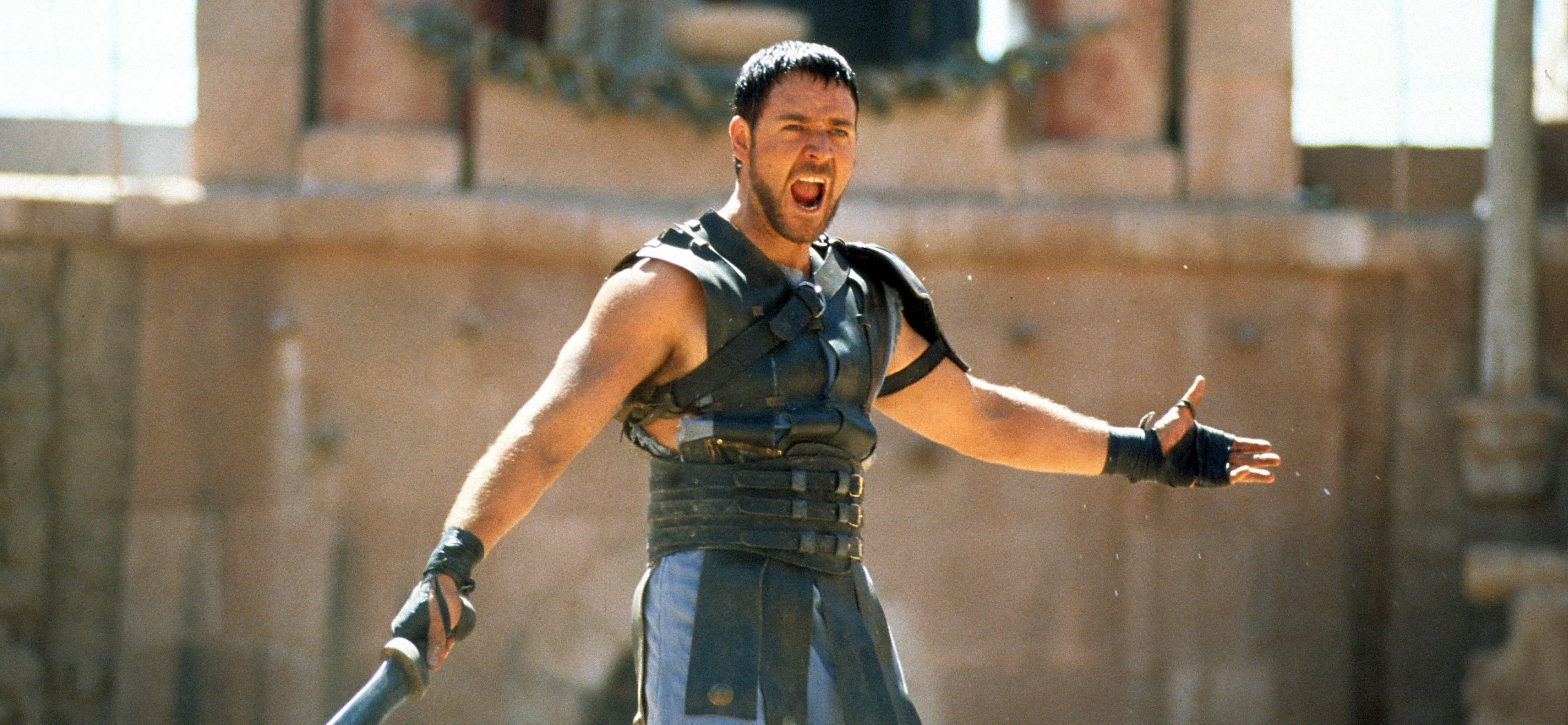 Russell Crowe in a gladiator outfit stands in an ancient arena, holding a sword and passionately shouting
