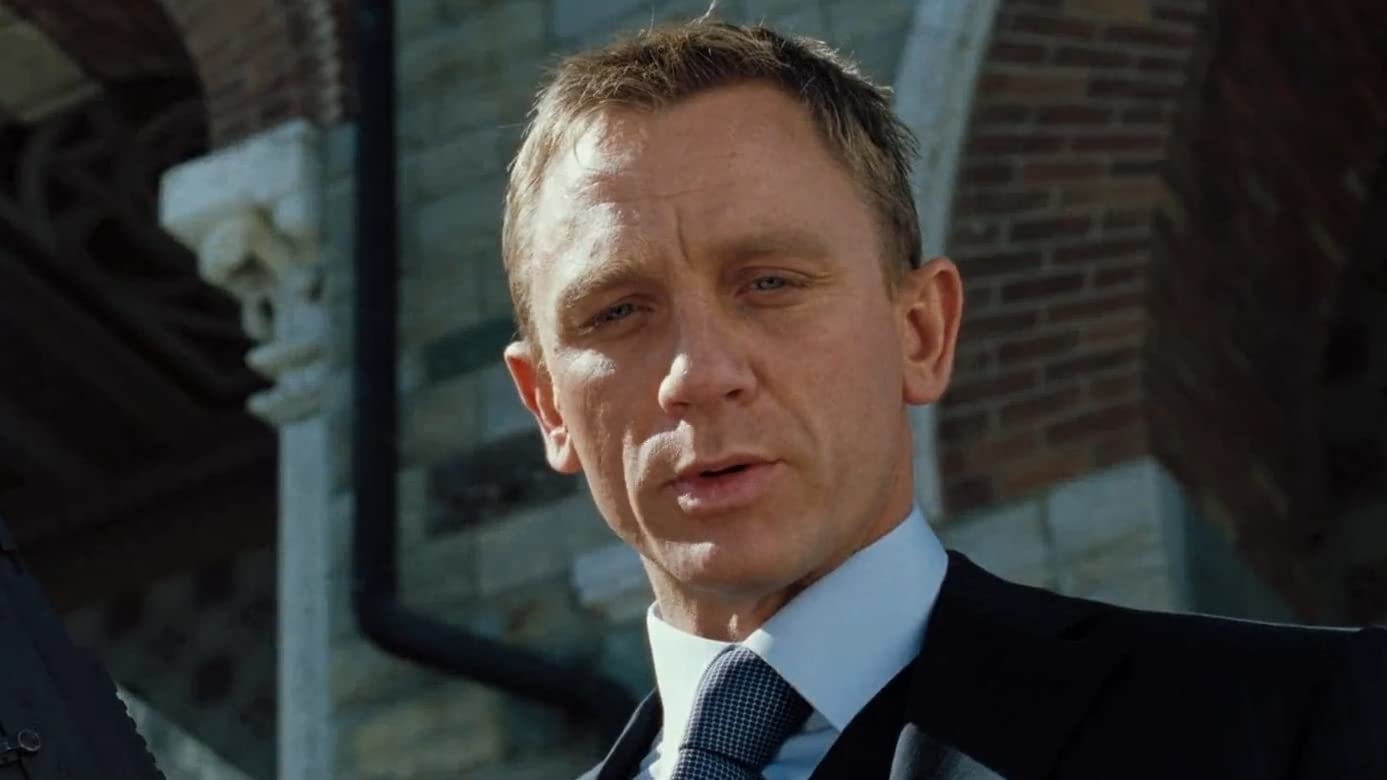 Daniel Craig is seen in a close-up shot, wearing a suit and tie, with a brick and stone building in the background