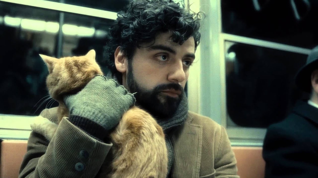 Man with a curly hair and beard, wearing a scarf and gloves, holding an orange cat in his arms on a subway train