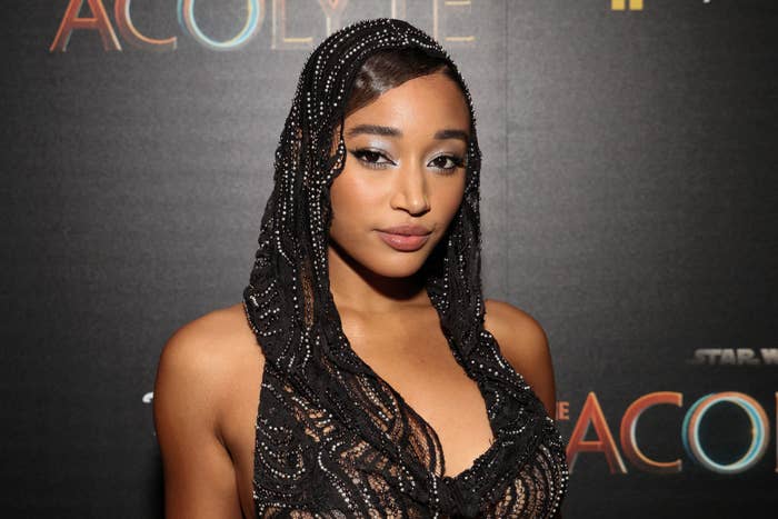 Amandla Stenberg at "The Acolyte" event, wearing a sequined outfit with a hood, posing for a photo in front of a branded backdrop