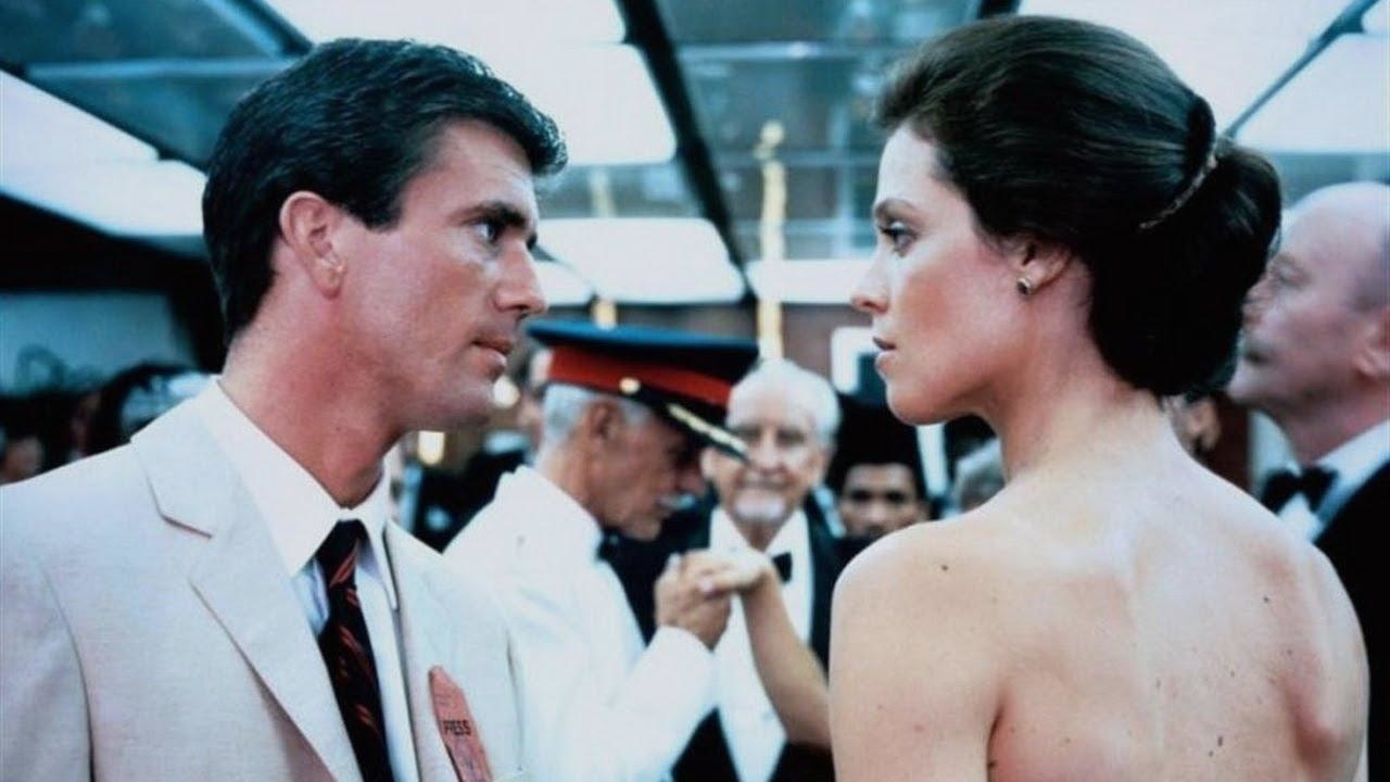 Mel Gibson and Sigourney Weaver in a formal event scene, having an intense conversation, others in background dressed in evening wear
