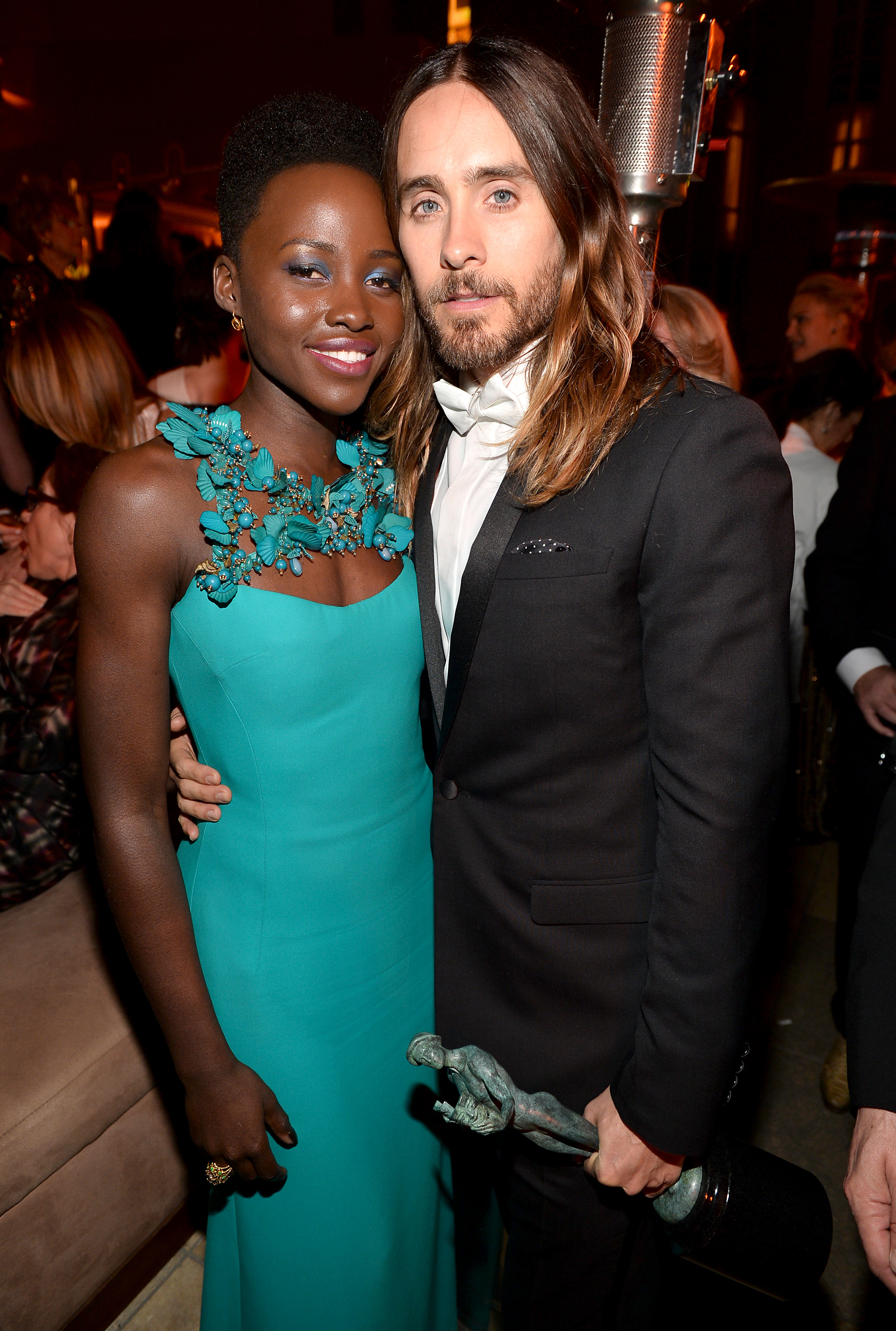 Lupita Nyong&#x27;o, wearing a gown with floral details, and Jared Leto in a suit, pose together at an event. Jared holds an award statue