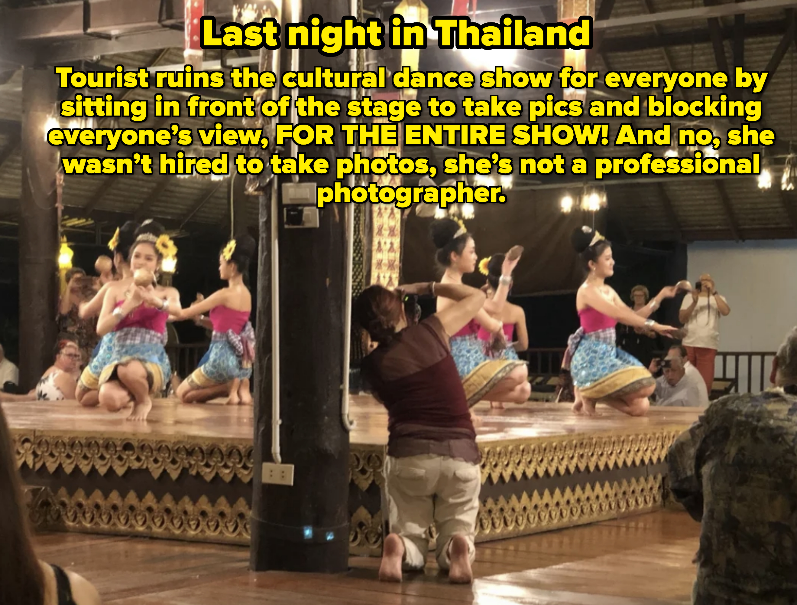 A group of dancers in traditional attire perform on a wooden stage while an audience member photographs them at an indoor event