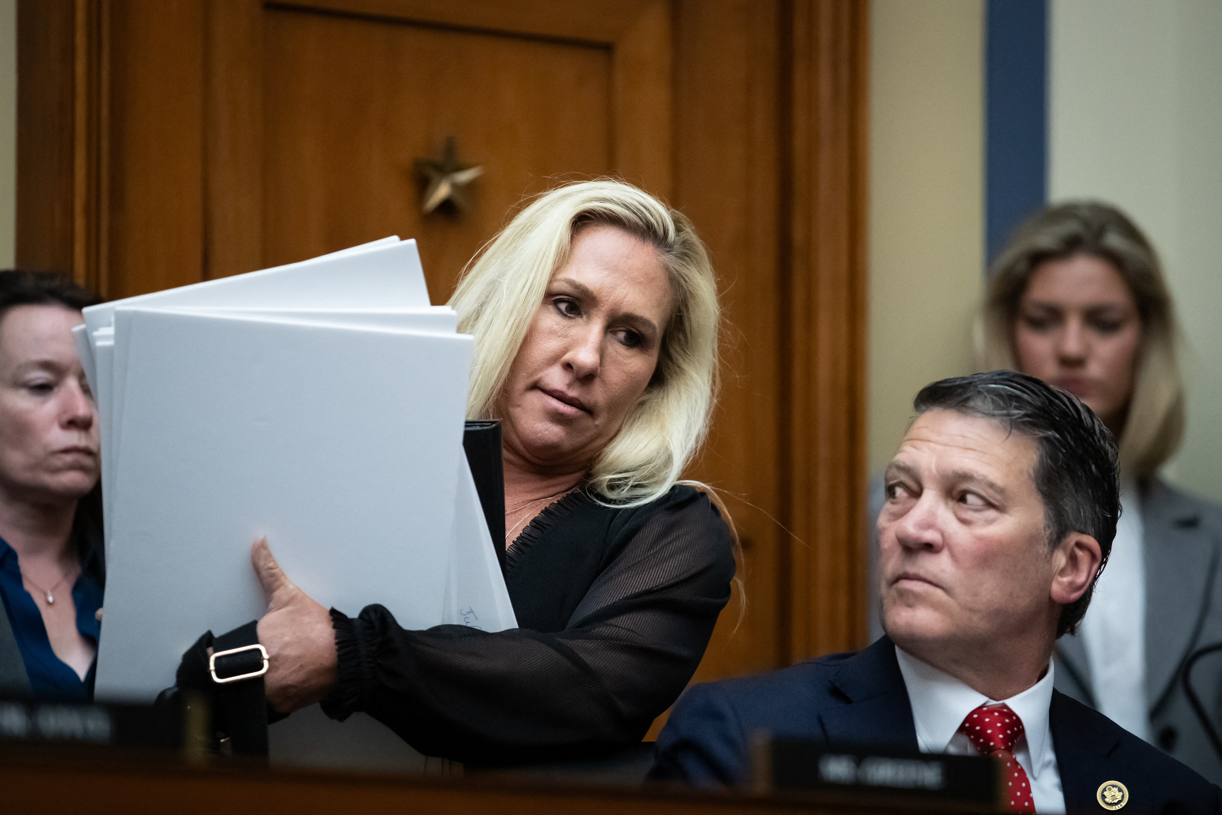 Marjorie Taylor Greene holds large papers and stands next to Mark E. Green, who sits at a desk. Two unidentified people are in the background