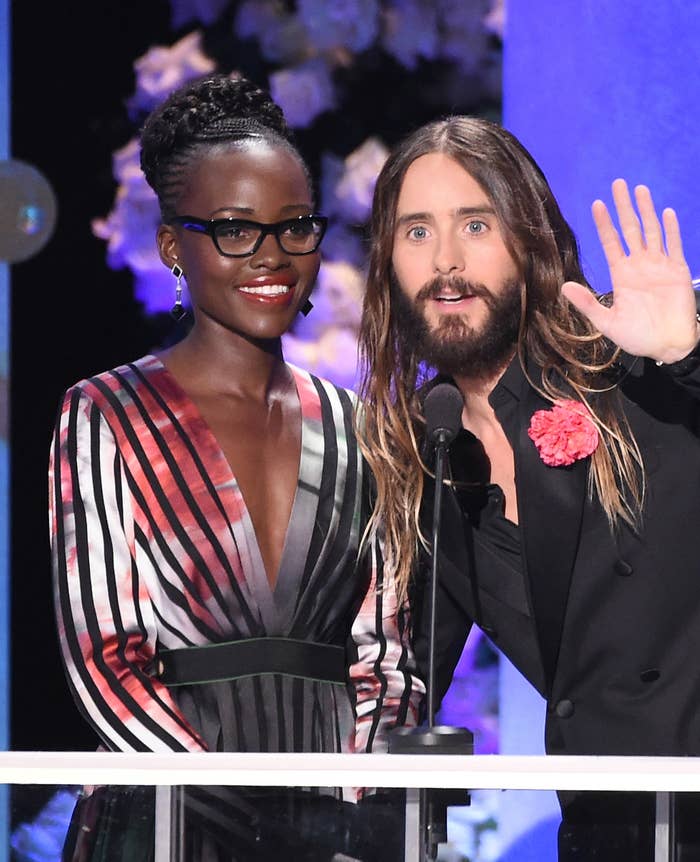 Lupita Nyong'o in a striped dress and glasses stands next to Jared Leto who's wearing a dark suit with a flower
