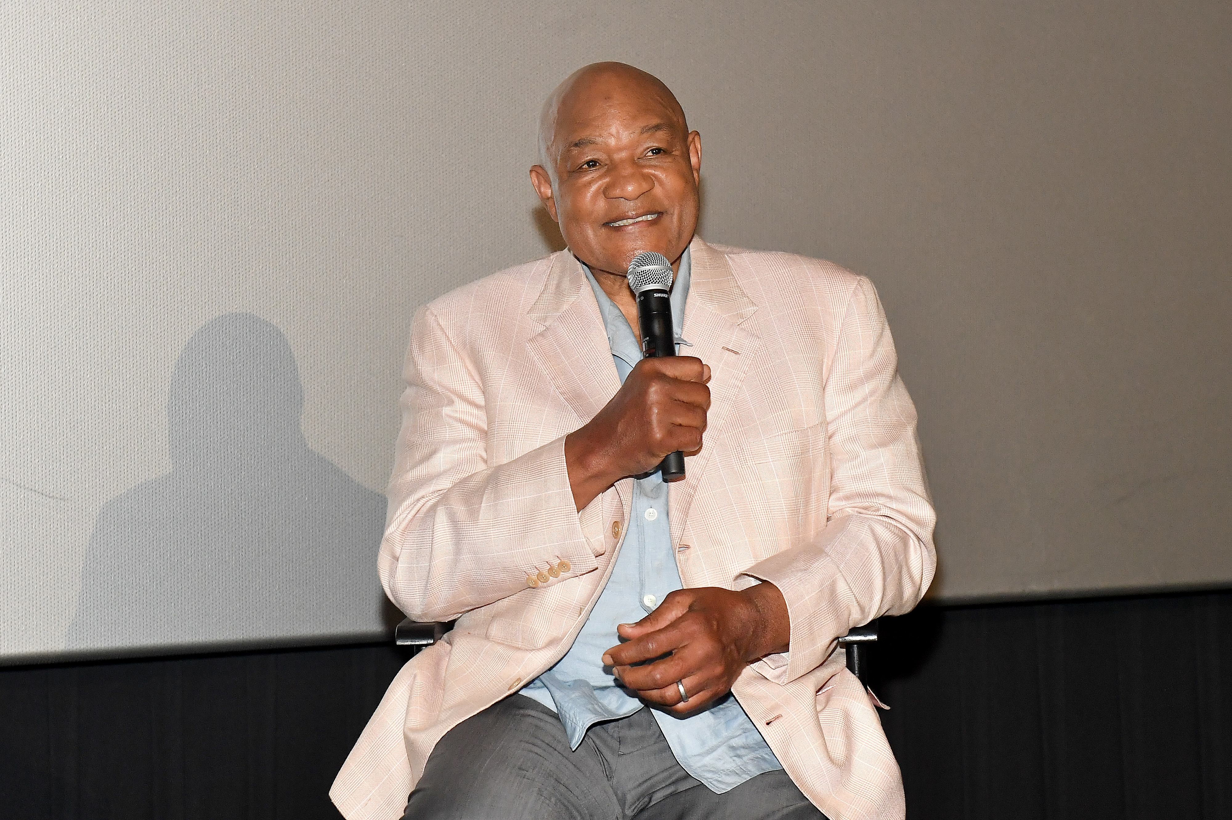 George Foreman speaking into a microphone, wearing a light blazer and sitting on stage