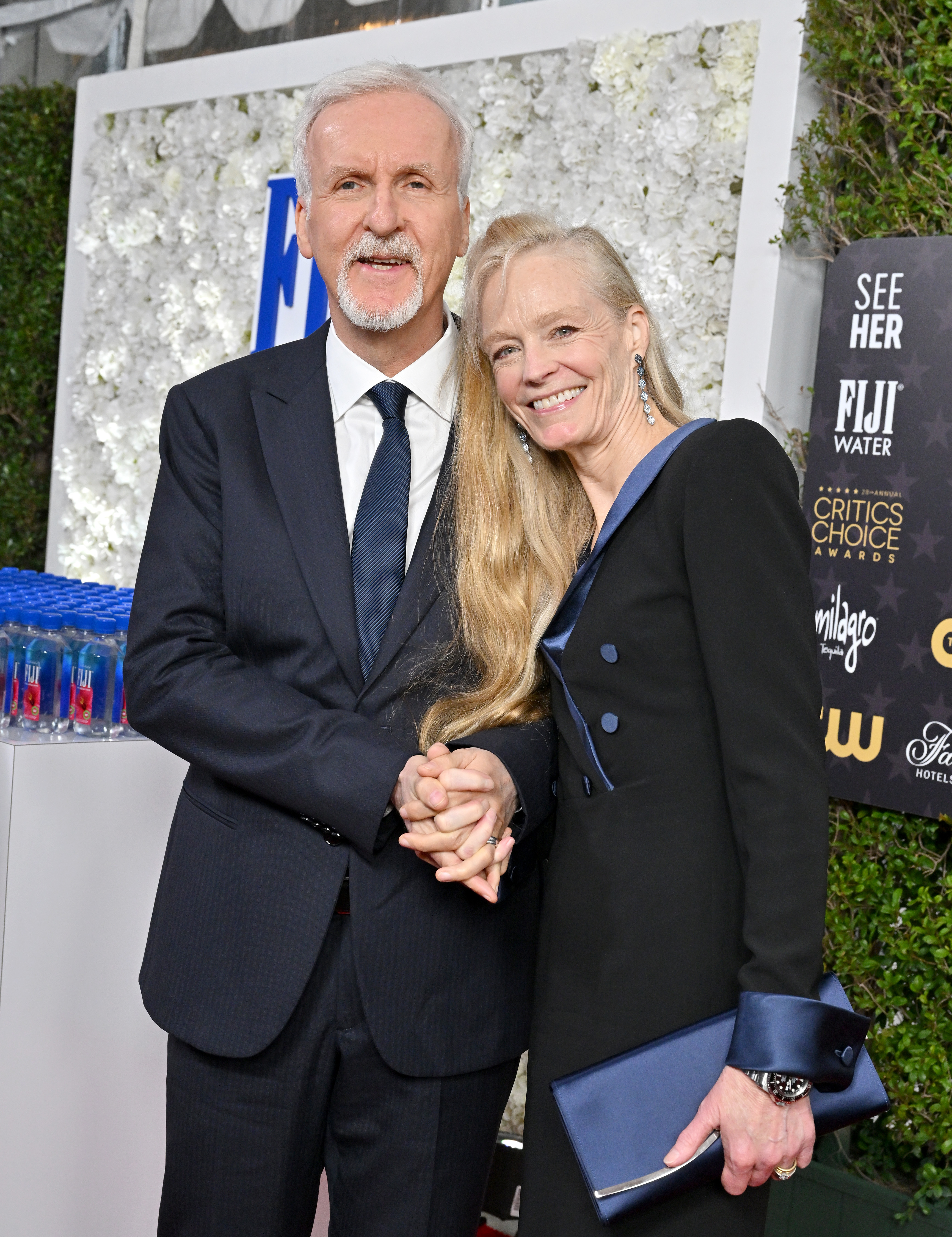 James Cameron and Suzy Amis Cameron hold hands on the red carpet. James wears a dark suit, while Suzy wears a double-breasted dress
