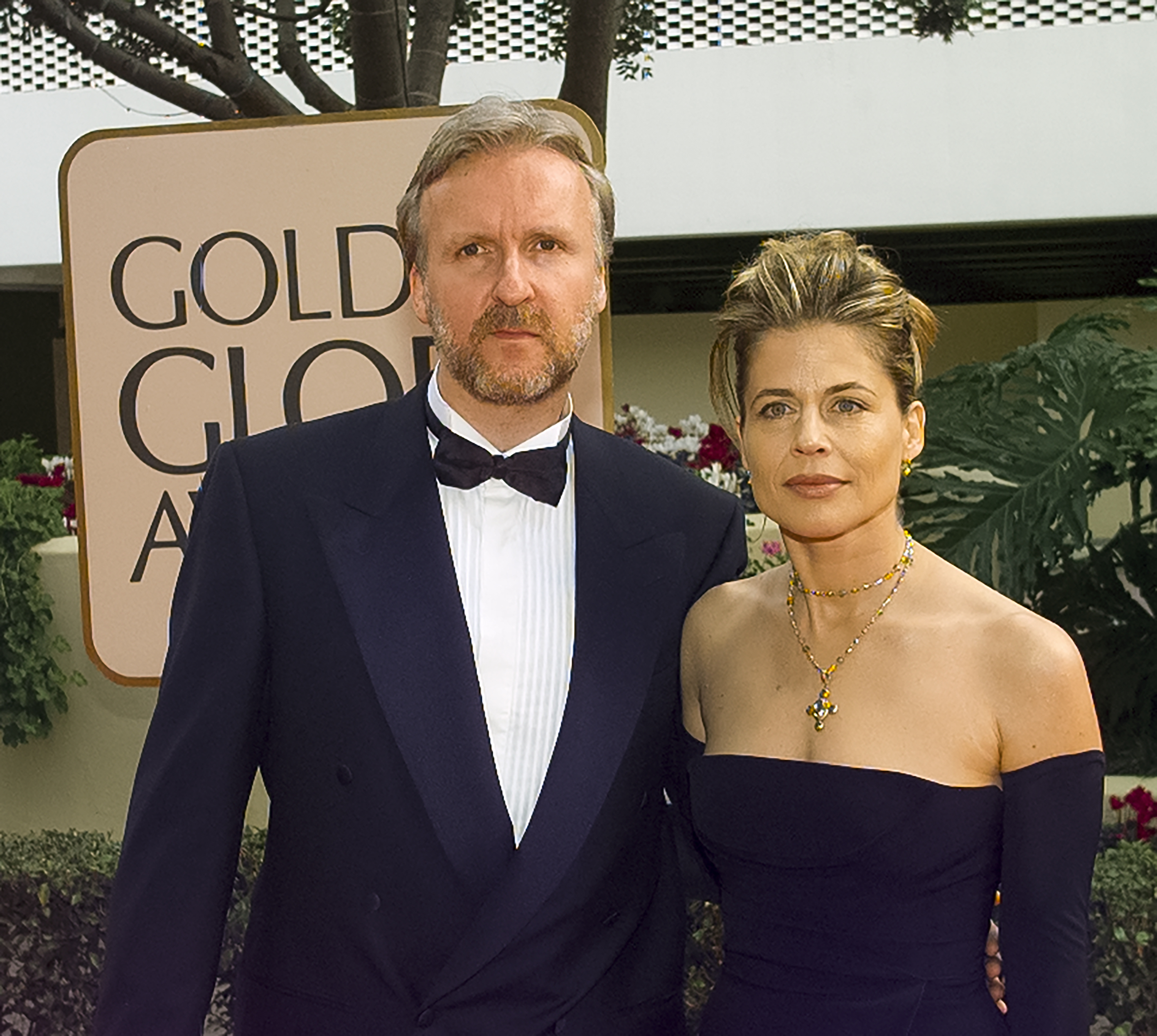 James Cameron and Linda Hamilton pose together at the Golden Globe Awards. James wears a black tuxedo, and Linda wears an elegant black, strapless gown