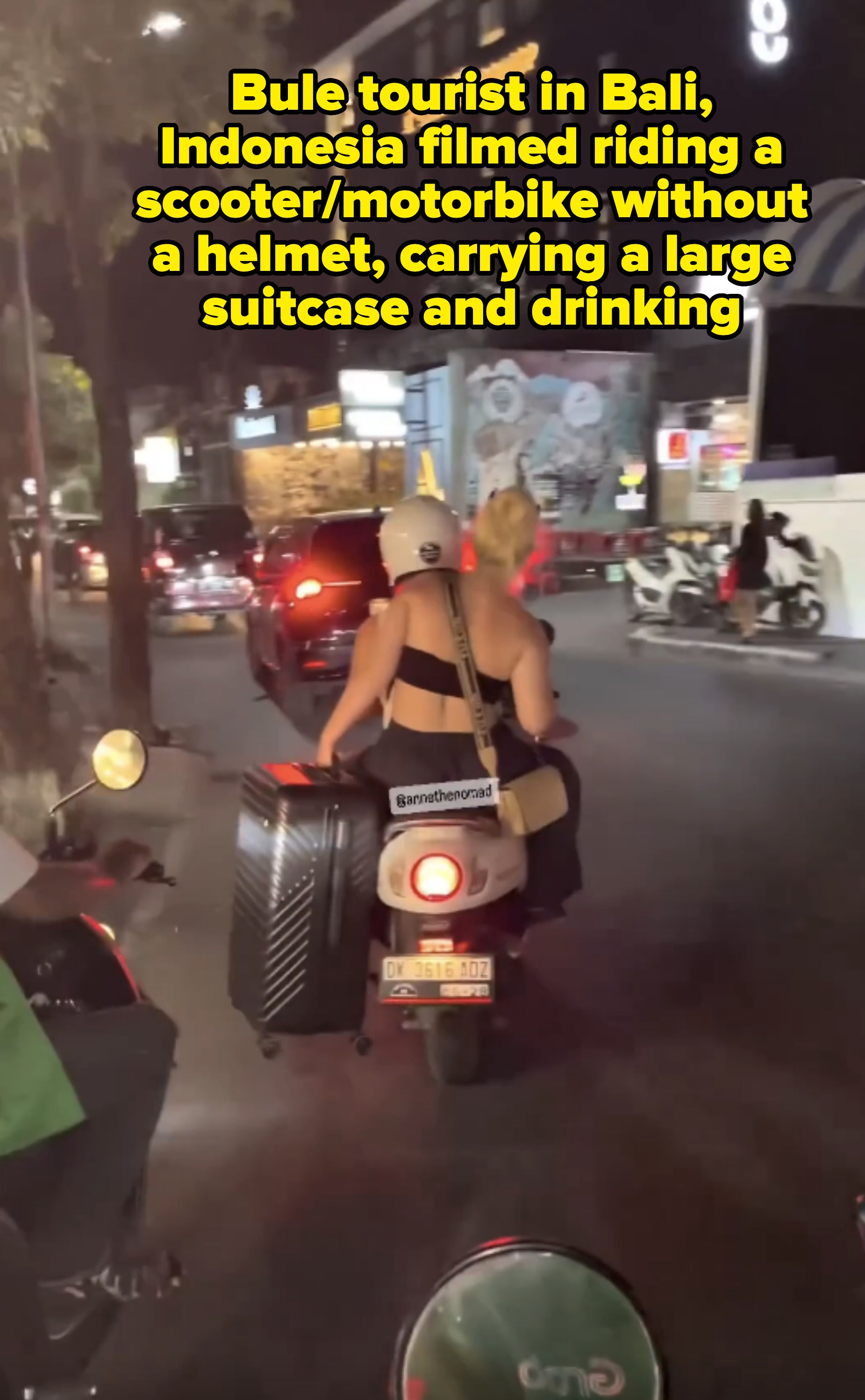 A woman on a motor scooter carries a large suitcase and a shoulder bag while riding through a busy city street at night