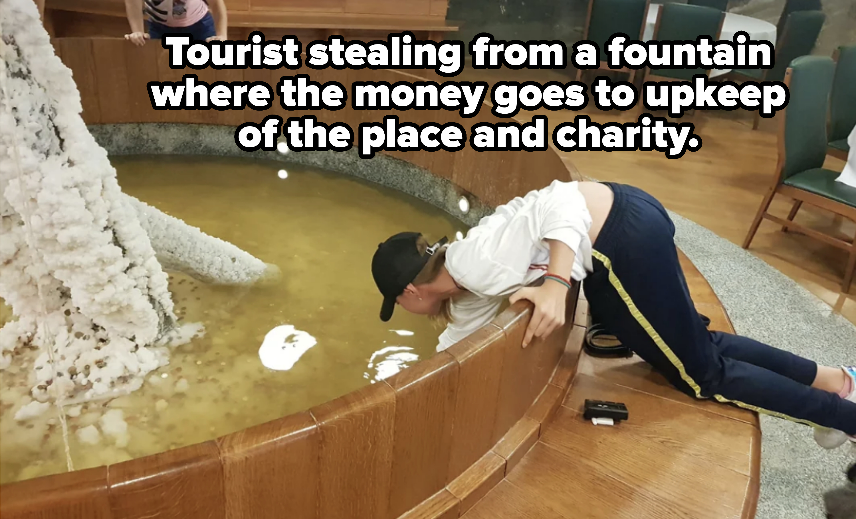 A person bends over a wooden railing to reach into a pool inside a public area, while another person stands nearby in the background