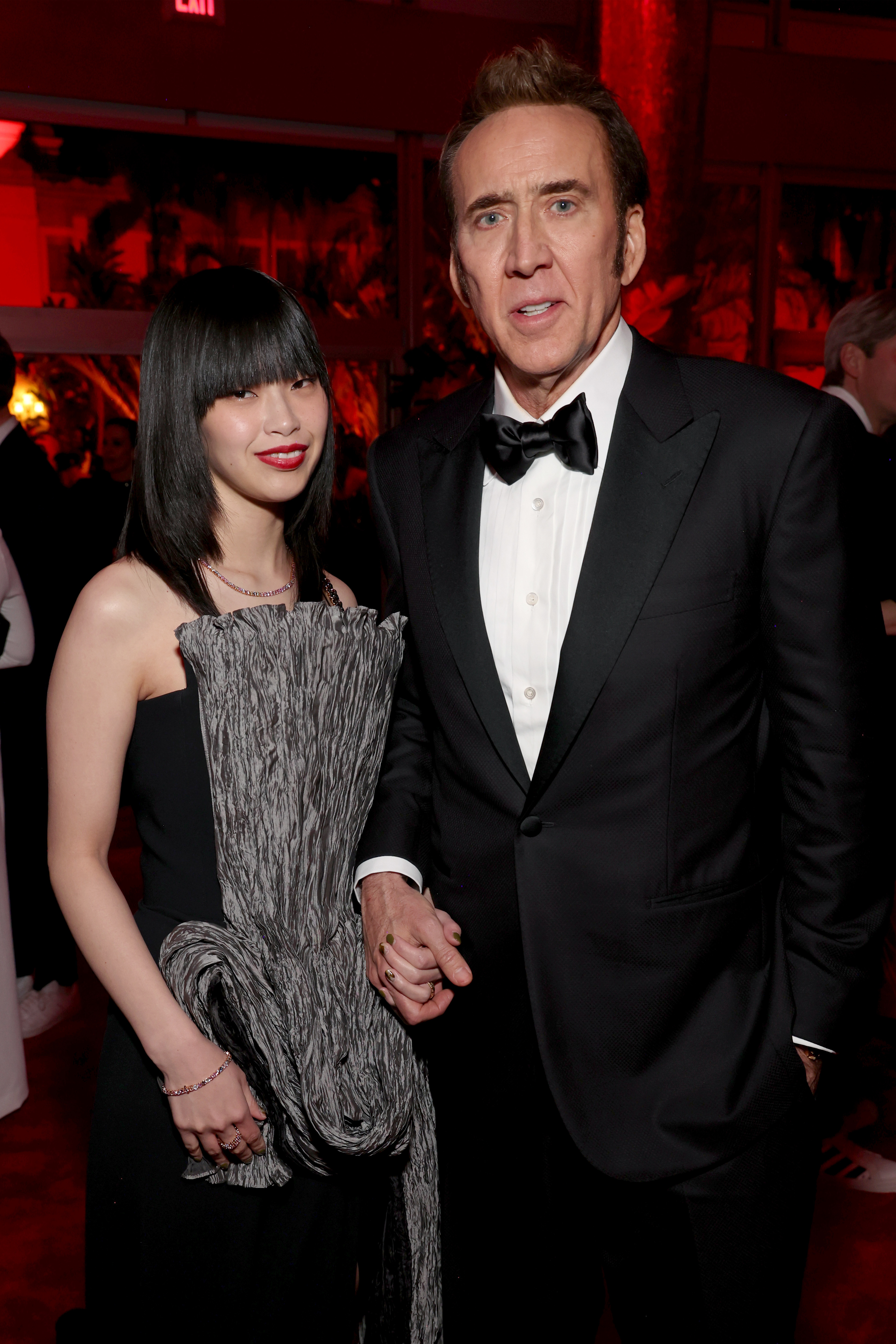 Nicolas Cage, in a classic black tuxedo, and Riko Shibata, in a unique black dress with textured detailing, pose together at a formal celebrity event