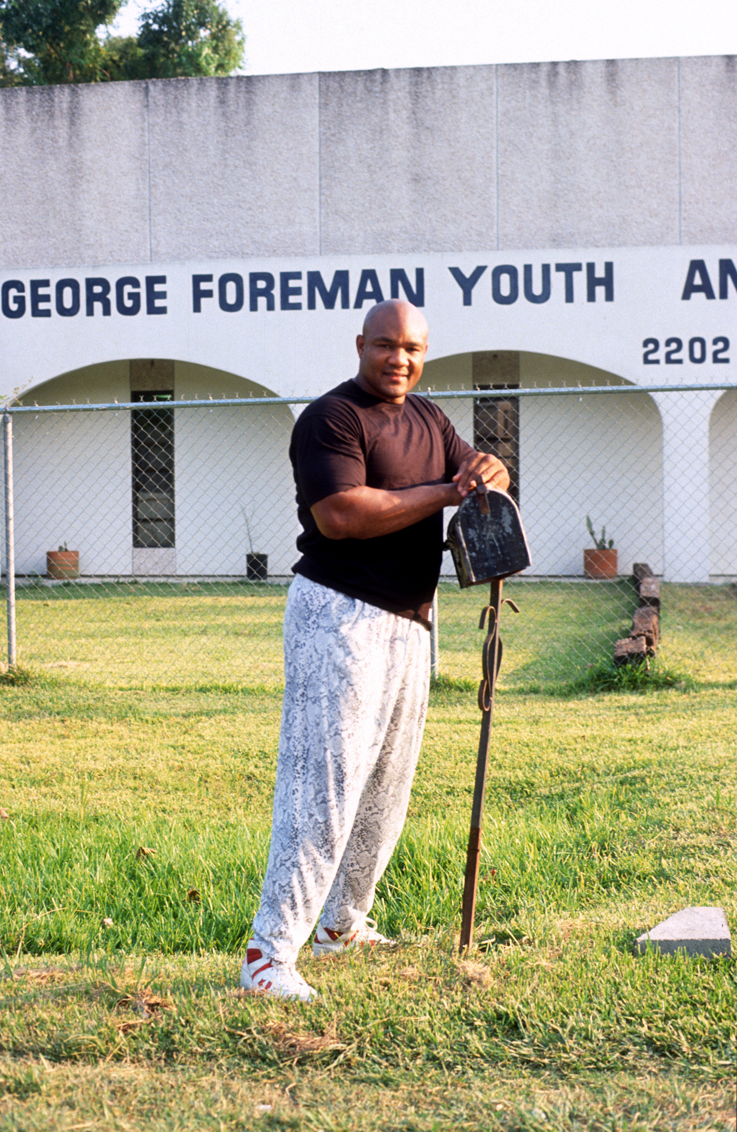 George Foreman holding a shovel and smiling in front of the George Foreman Youth Center sign. He is wearing a black shirt and patterned pants