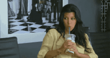 Kim Kardashian drinks from a straw cup, looking to her side in a distracted manner while seated