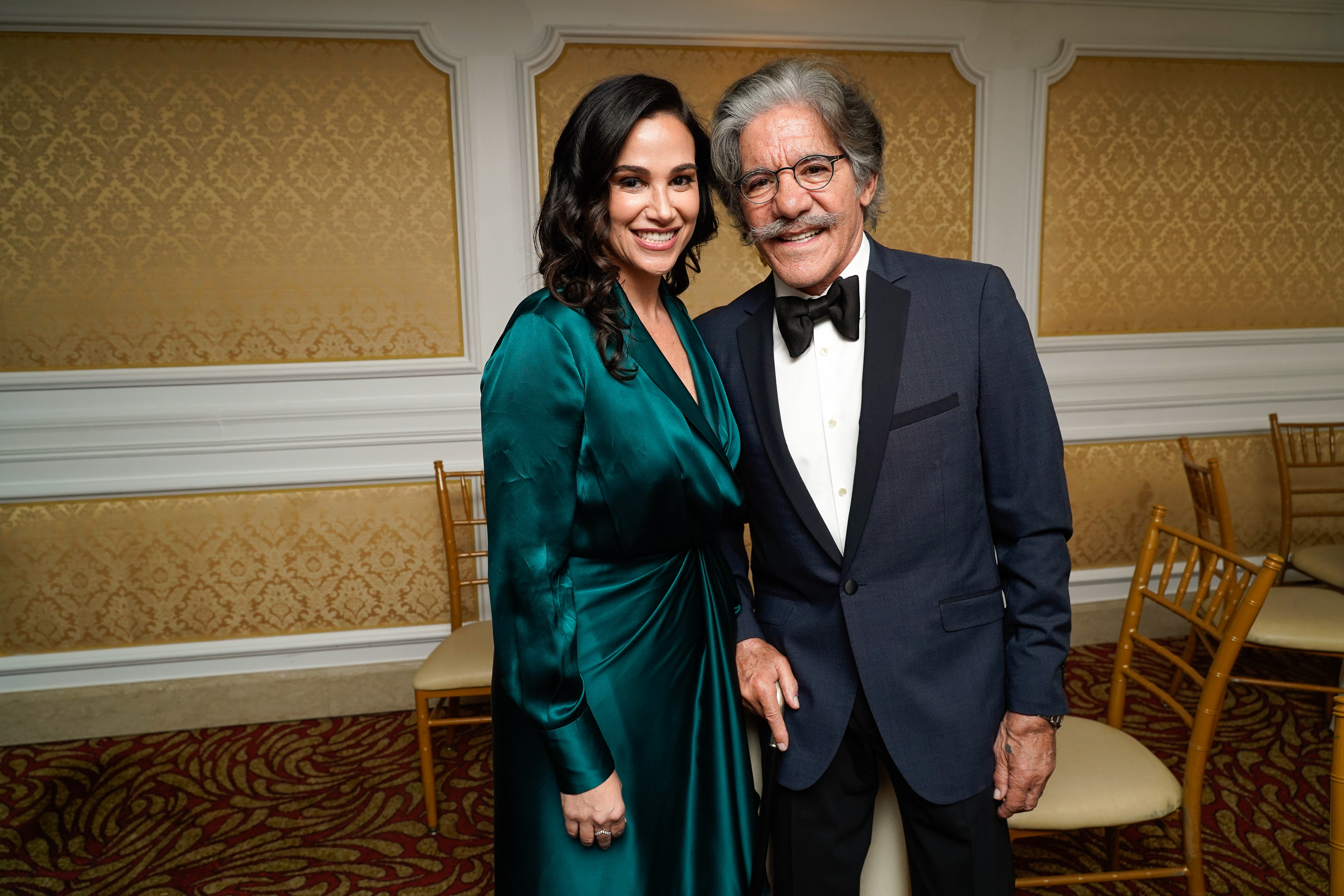 Smiling Geraldo Rivera in a black suit and bowtie standing next to Erica Michelle Levy in an elegant, long-sleeve, satin dress at an indoor event