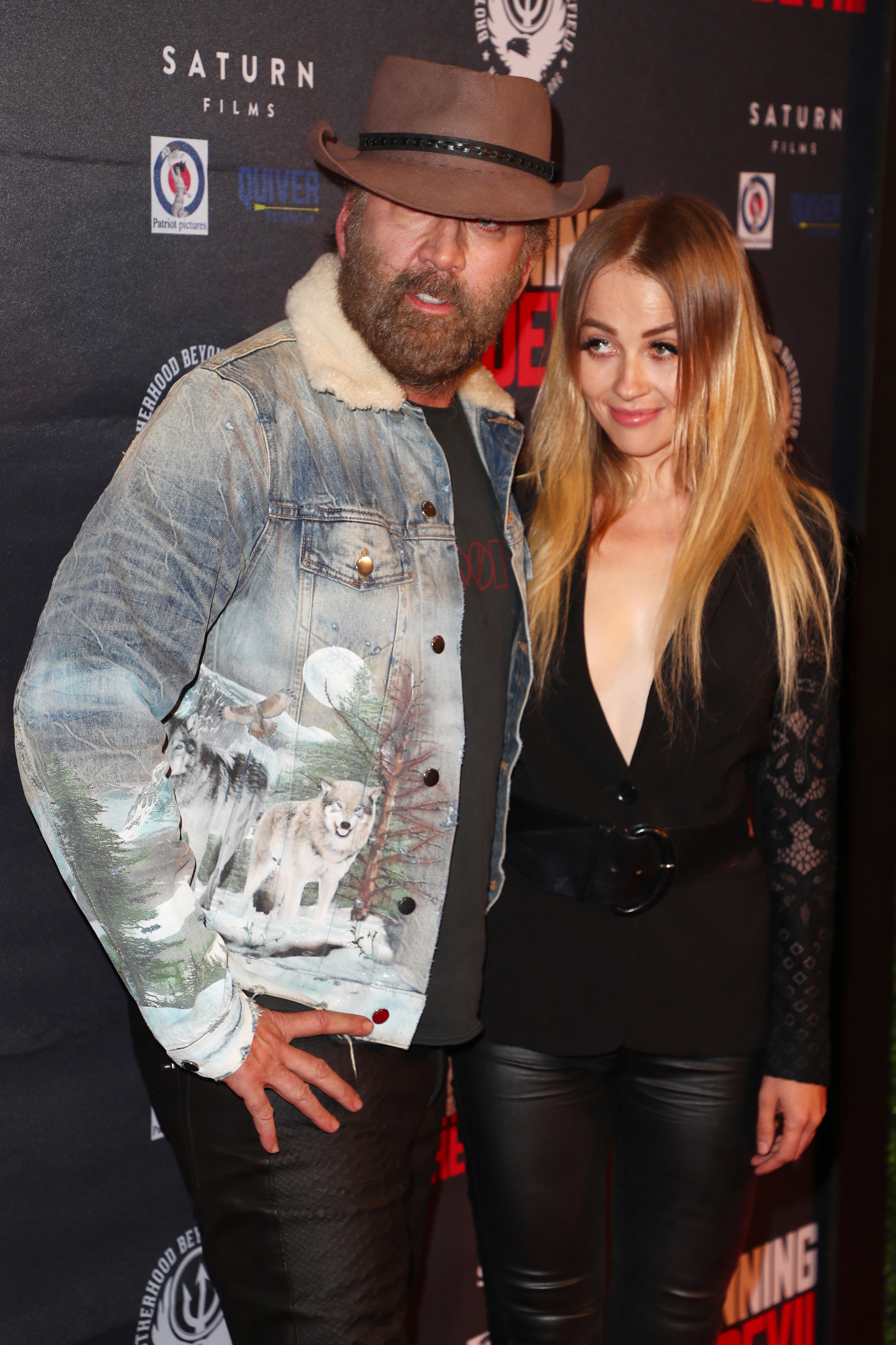 Nicolas Cage and Erika Koike on the red carpet. Nicolas wears a denim jacket with a wolf design and a brown hat, while Christina wears a plunging black outfit