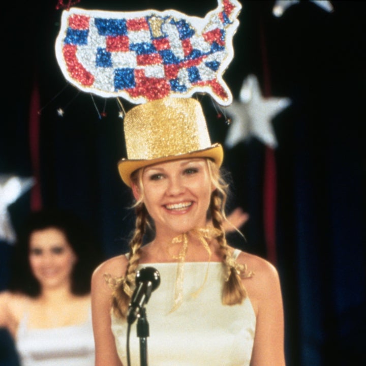 Kirsten Dunst stands on stage wearing a light dress and a sequined USA hat, with a large microphone in front of her. Another woman with number 7 is seen in the background