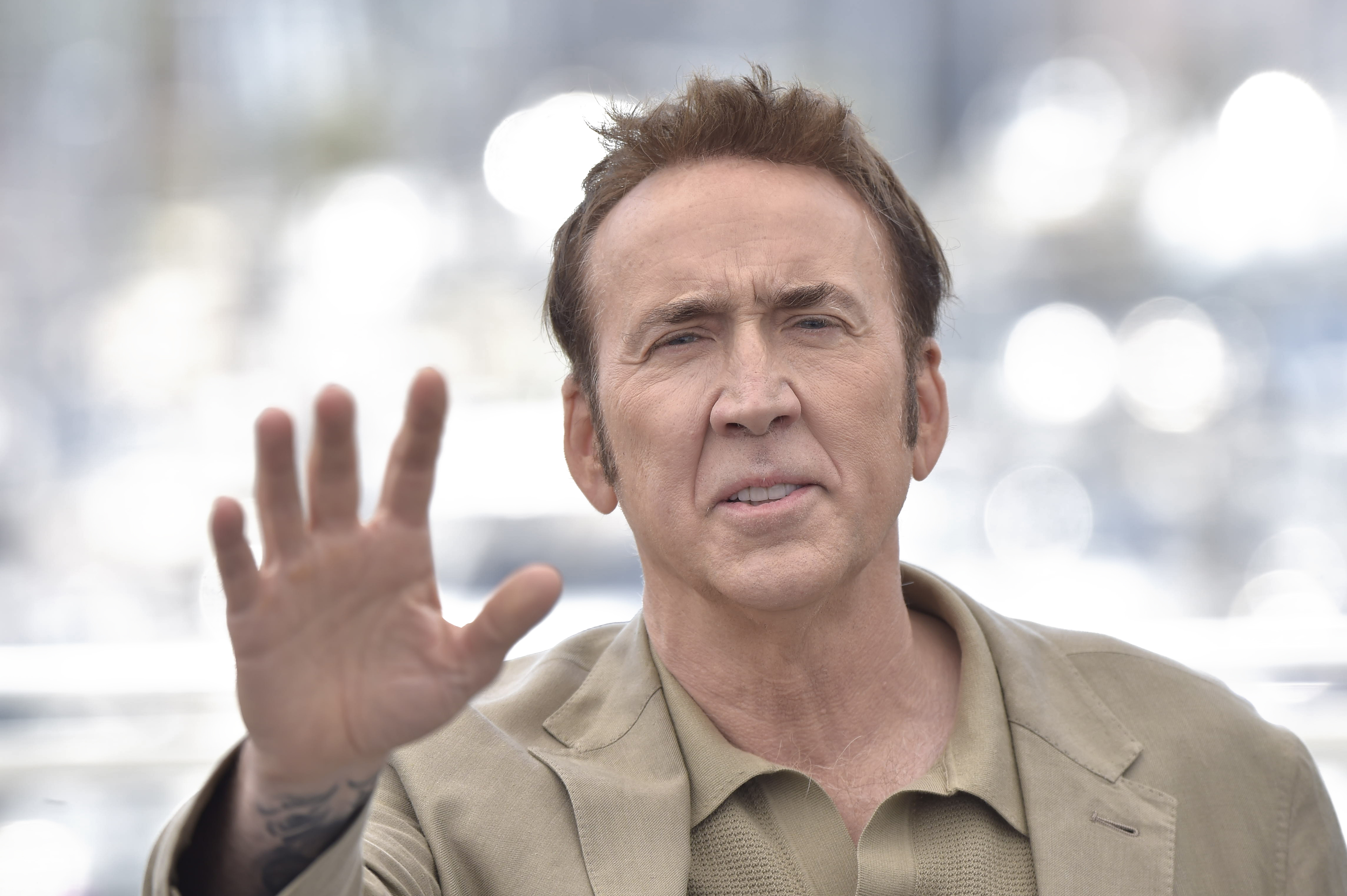 Nicolas Cage raises his hand in a wave while wearing a light-colored jacket and shirt during a public appearance