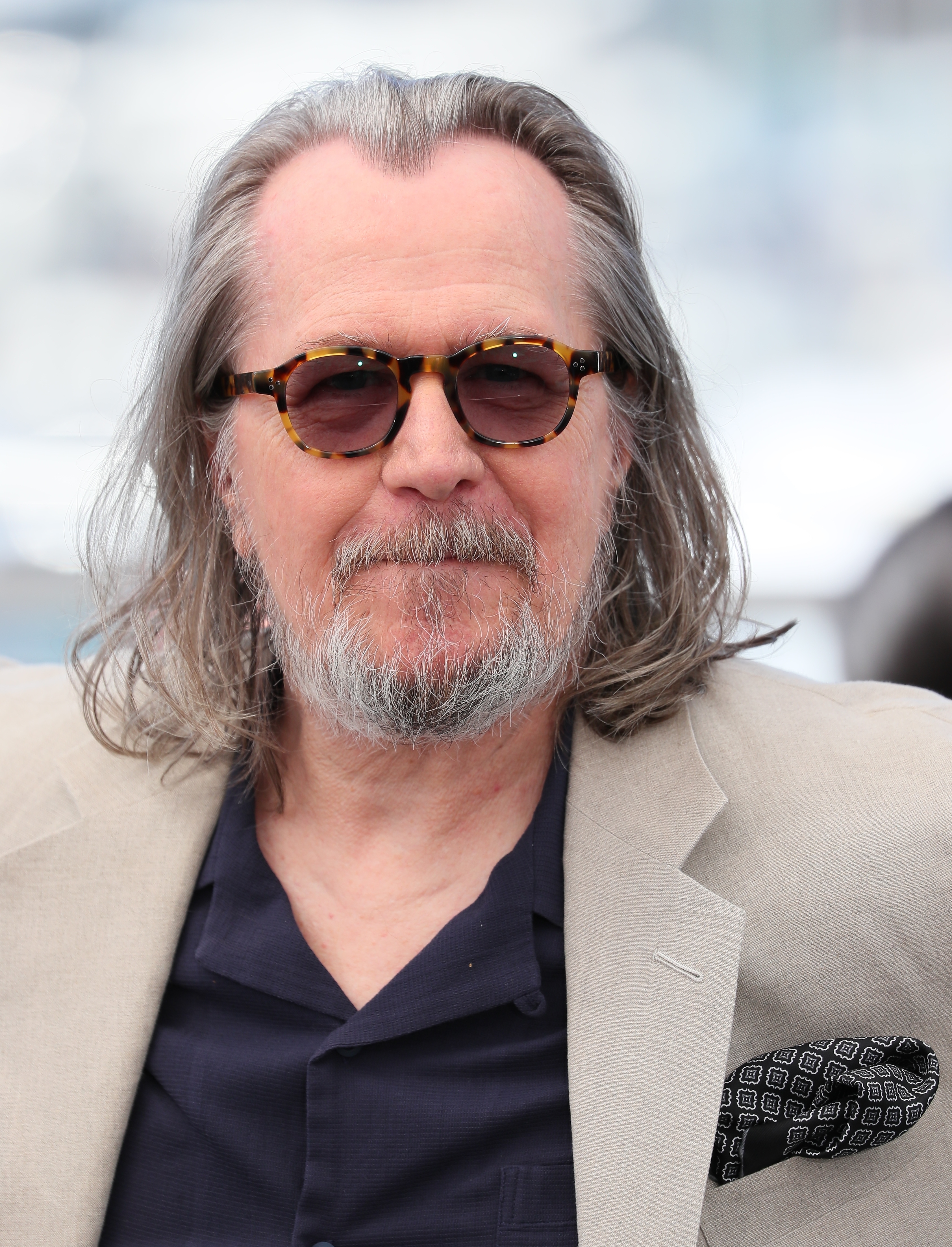 Gary Oldman wearing a light blazer over a dark shirt with sunglasses, posing for a photo