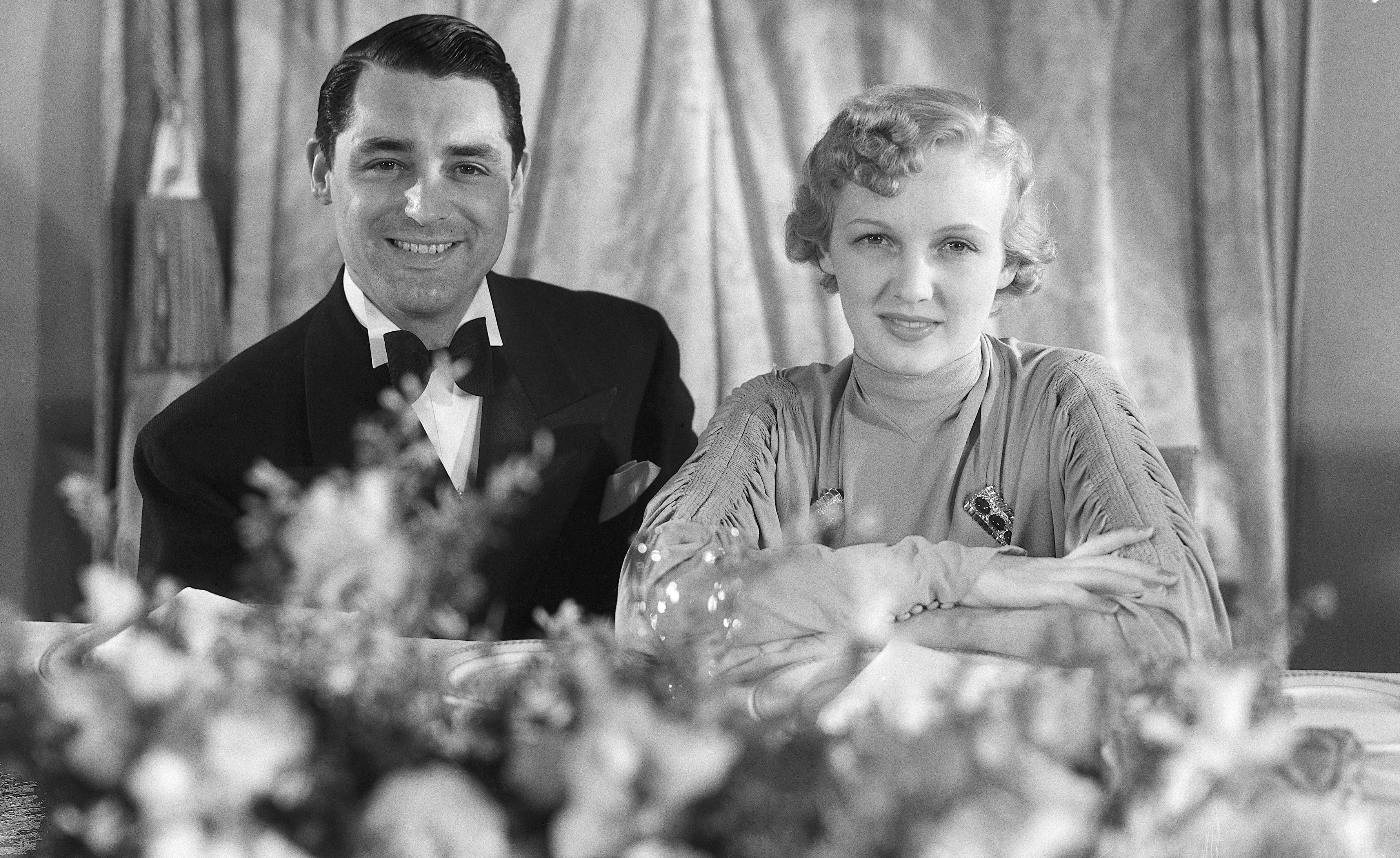 Cary Grant in a tuxedo and Mary Brian in an elegant gown are seated, smiling, and posing for a photo against a curtain backdrop