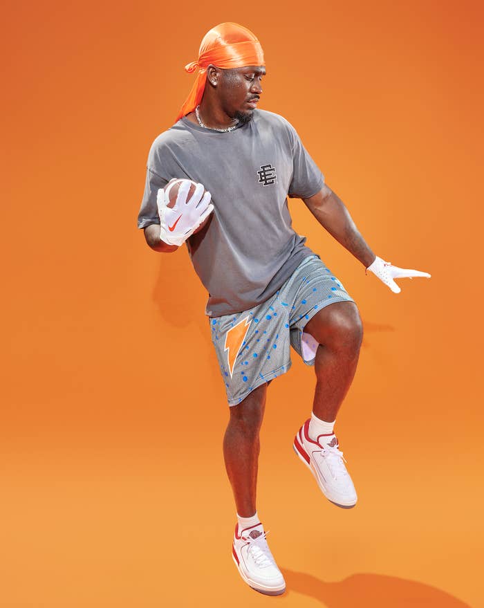 Man in a sporty outfit jumps mid-air. He wears a gray t-shirt, printed shorts, white sneakers, and an orange headscarf