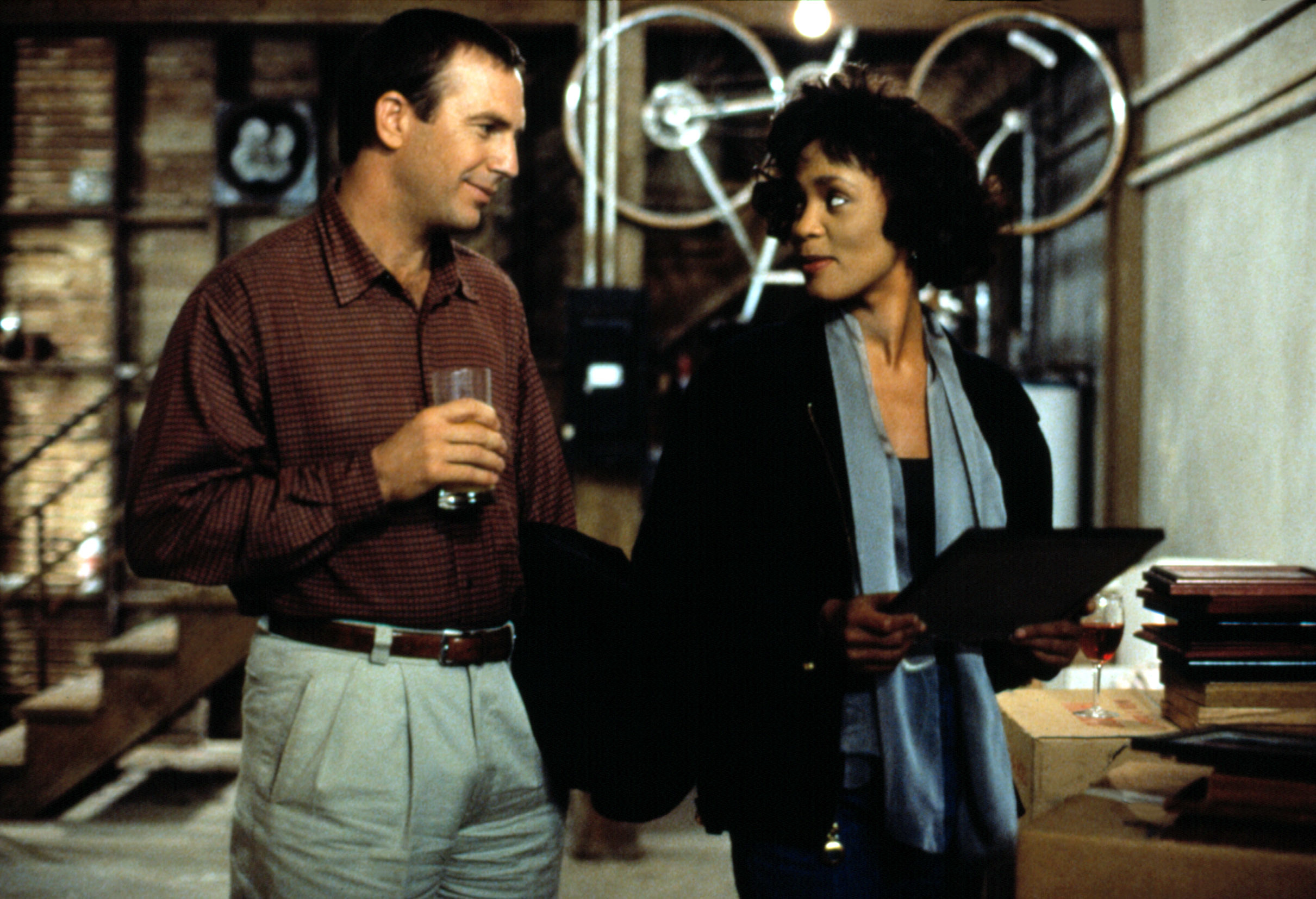 Kevin Costner and Whitney Houston in a film scene, with Costner holding a drink and Houston holding a folder, standing in a room with industrial decor