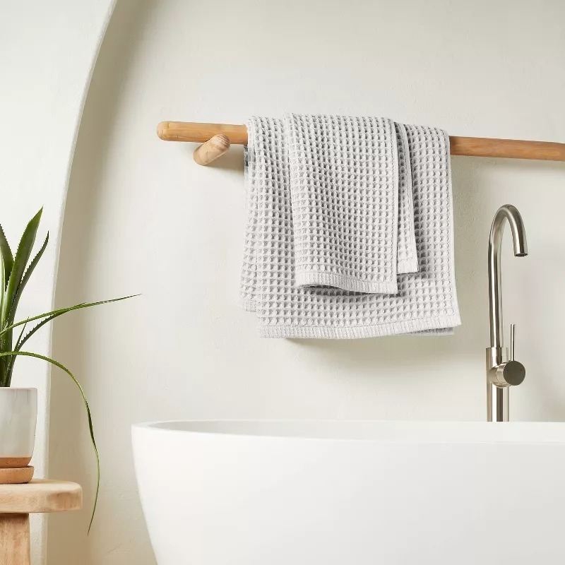A modern bathroom with a white freestanding bathtub, a sleek faucet, and light grey textured towels hanging on a wooden rod. A potted plant is placed on a wooden stool