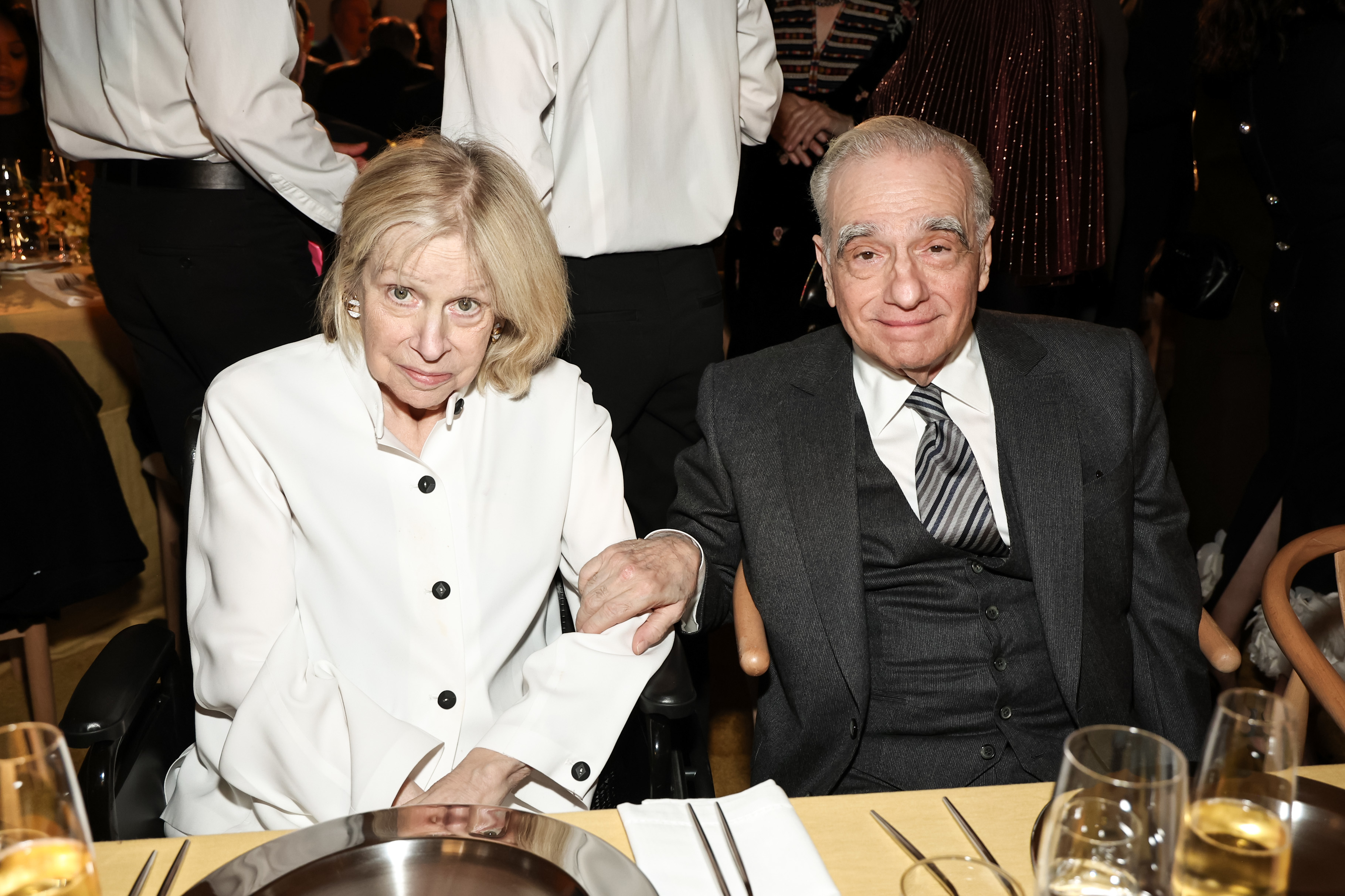 Two people seated at a dinner event, one in a white coat and the other in a suit and striped tie, holding hands over the table