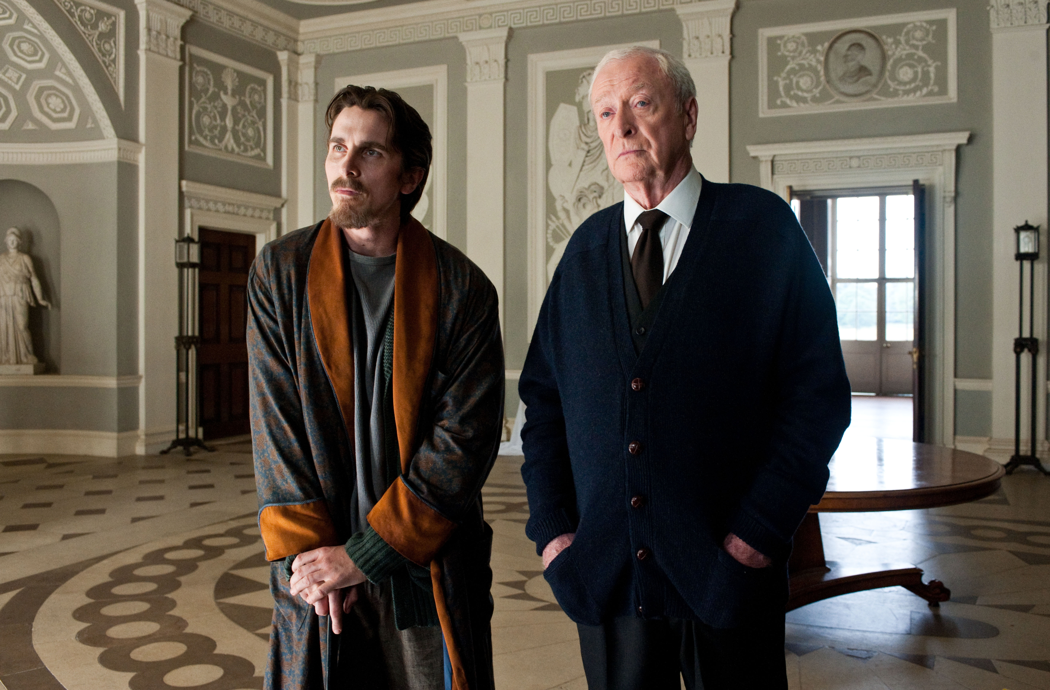 Christian Bale in a robe and Michael Caine in a cardigan stand in an ornate room with patterned floors and intricate wall designs