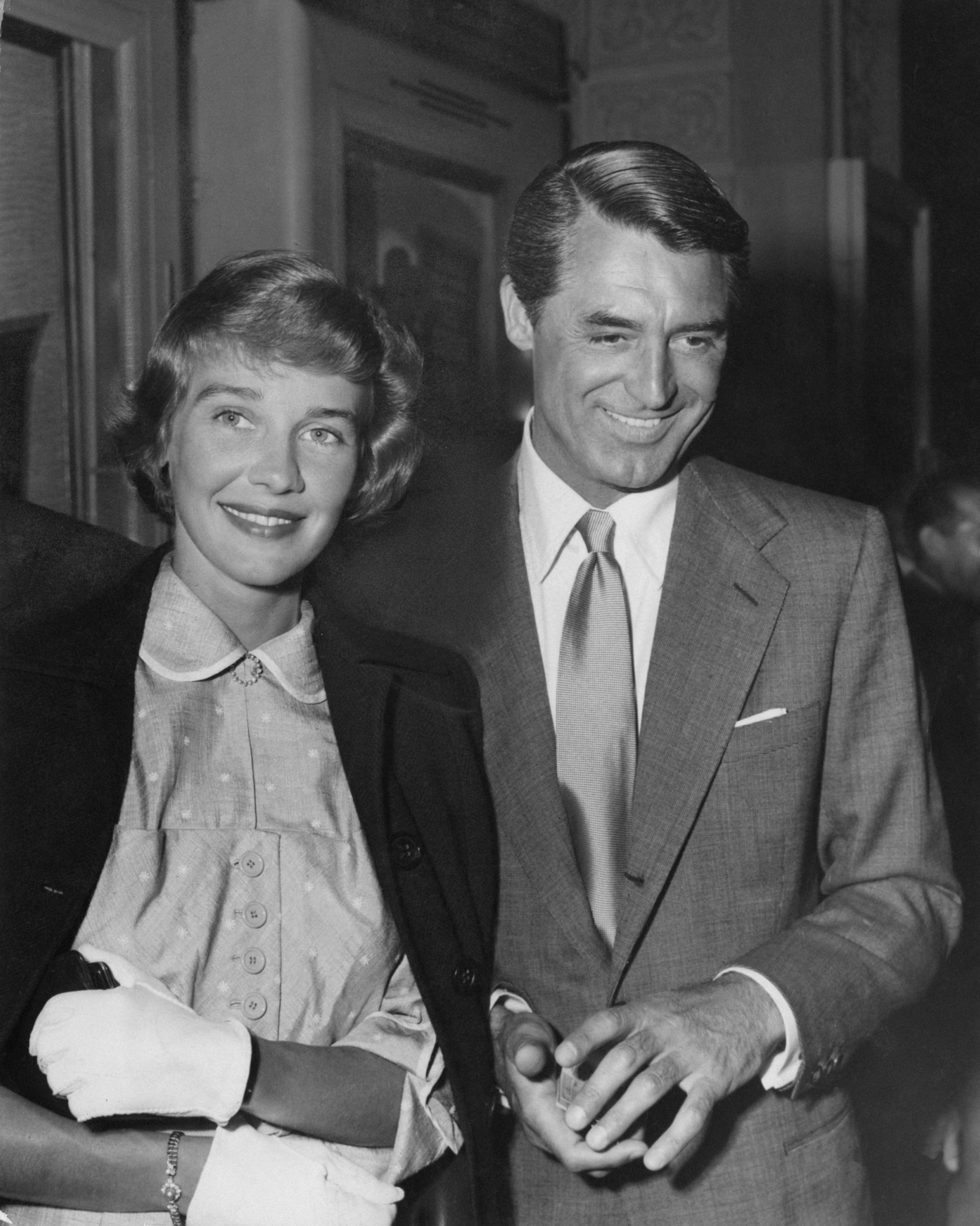 Cary Grant and Betsy Drake smiling, with Grant wearing a suit and tie and Drake in a collared dress and coat
