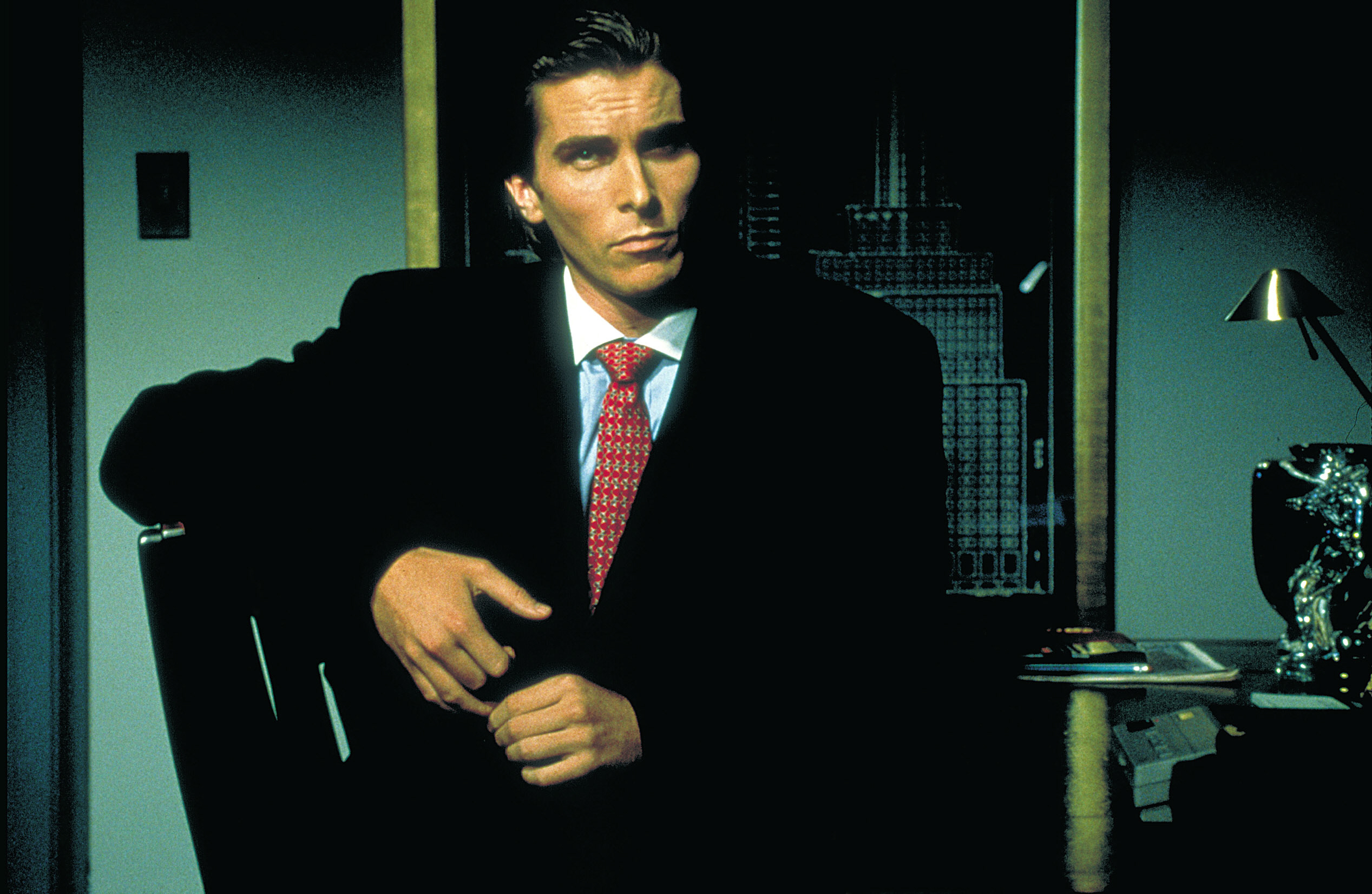 Christian Bale in a dark suit with a tie, posing confidently in an office setting in a scene from American Pyscho