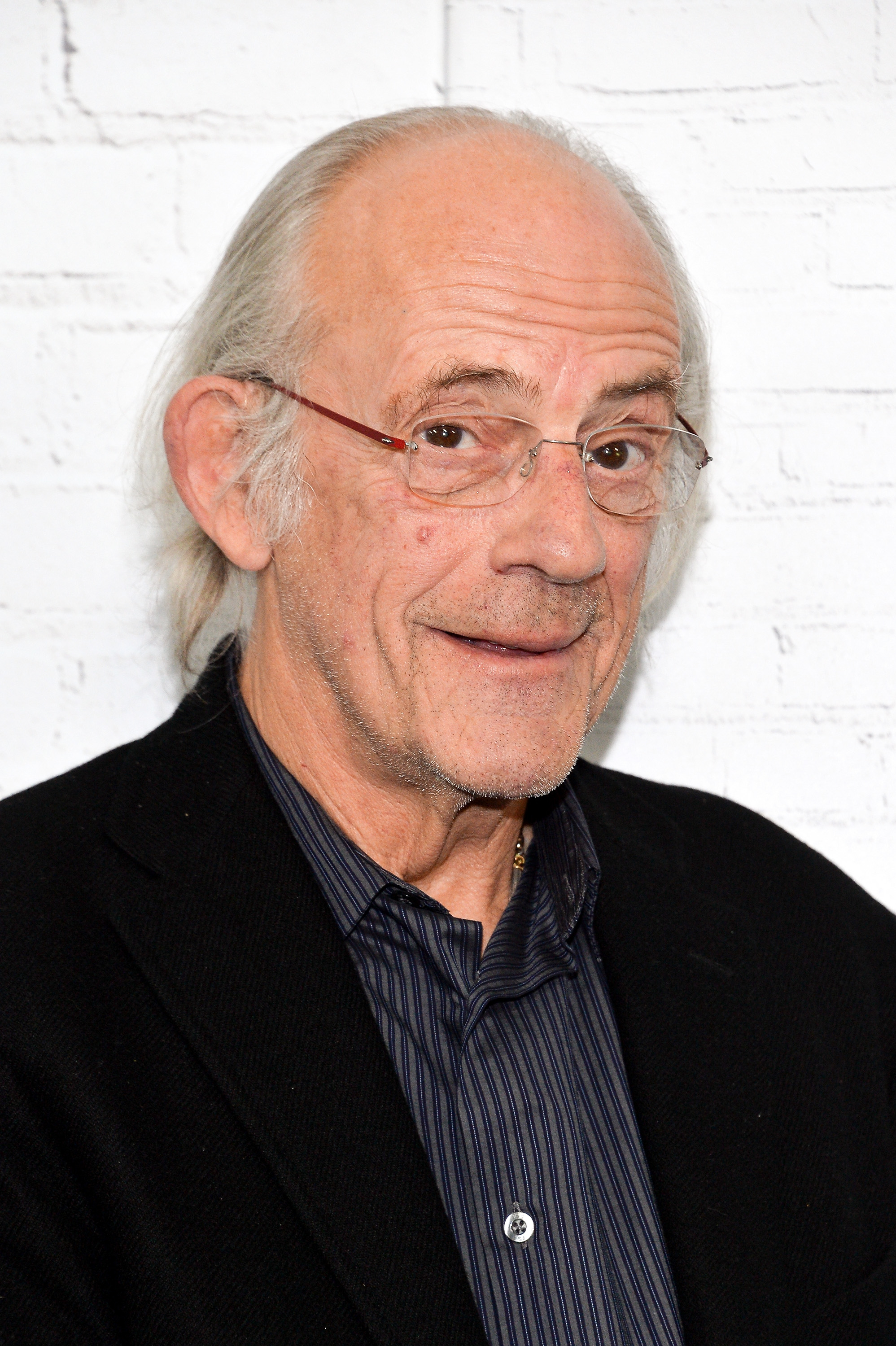 Christopher Lloyd smiling at the camera in a black blazer over a striped shirt, with a white brick wall in the background