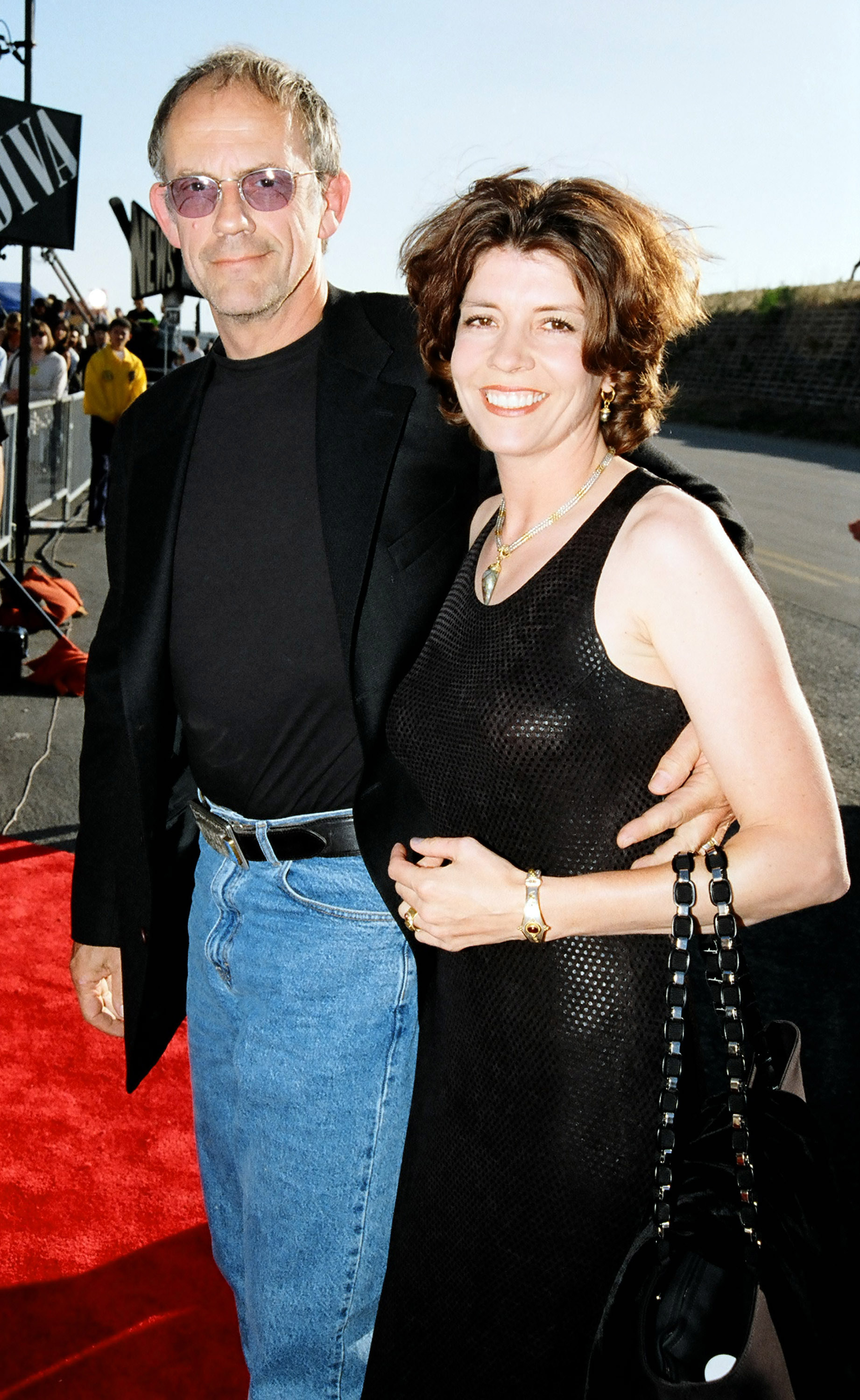 Man and woman smiling on a red carpet, man in black blazer and jeans, woman in black sleeveless dress, posing for the cameras