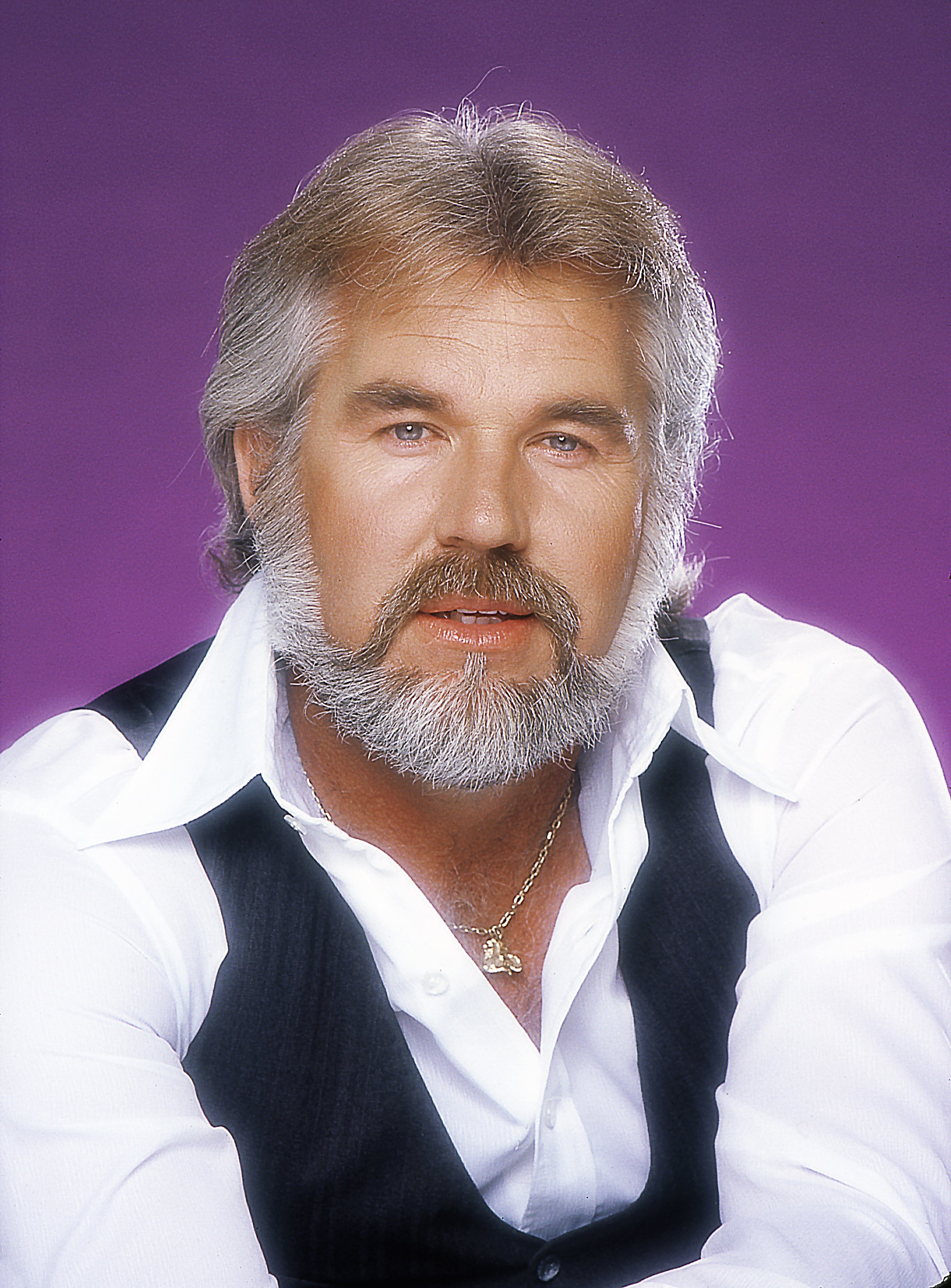 Kenny Rogers poses in a studio, wearing a white shirt, black vest, and gold chain