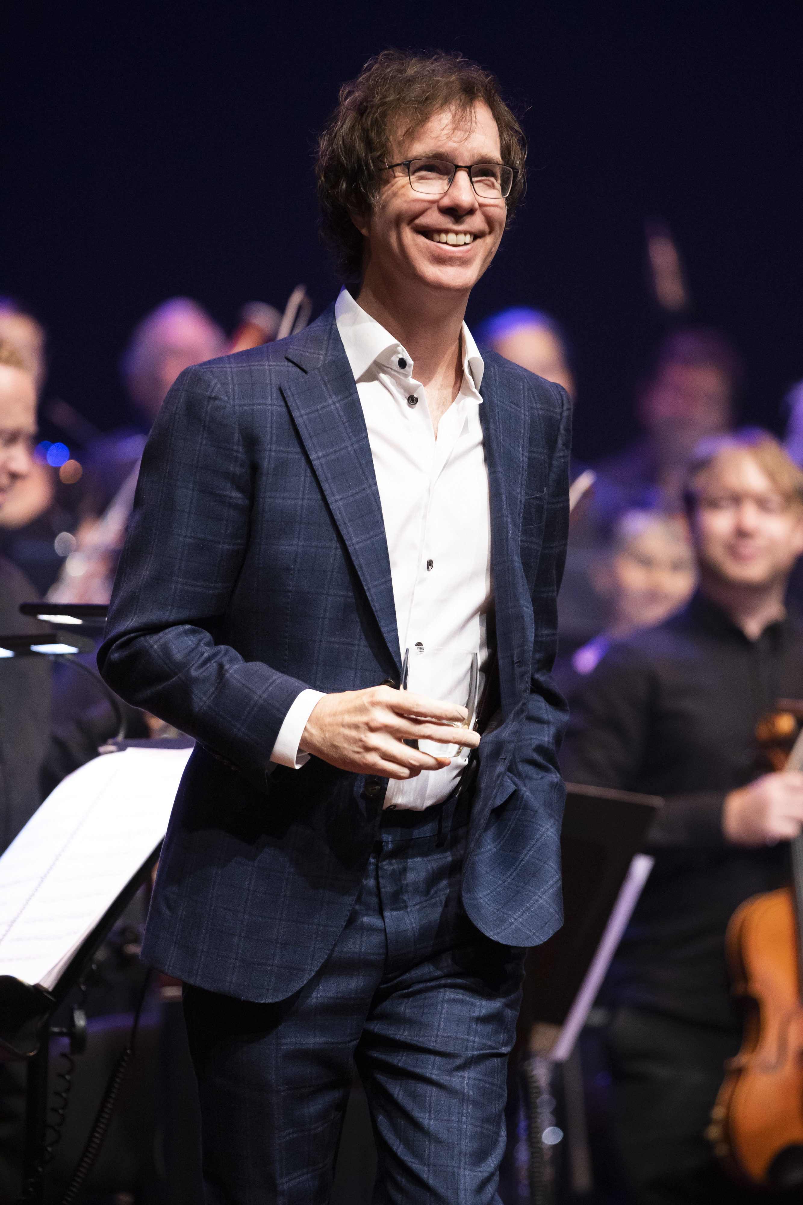 Ben Folds, wearing a patterned suit and glasses, performs on stage with an orchestra in the background