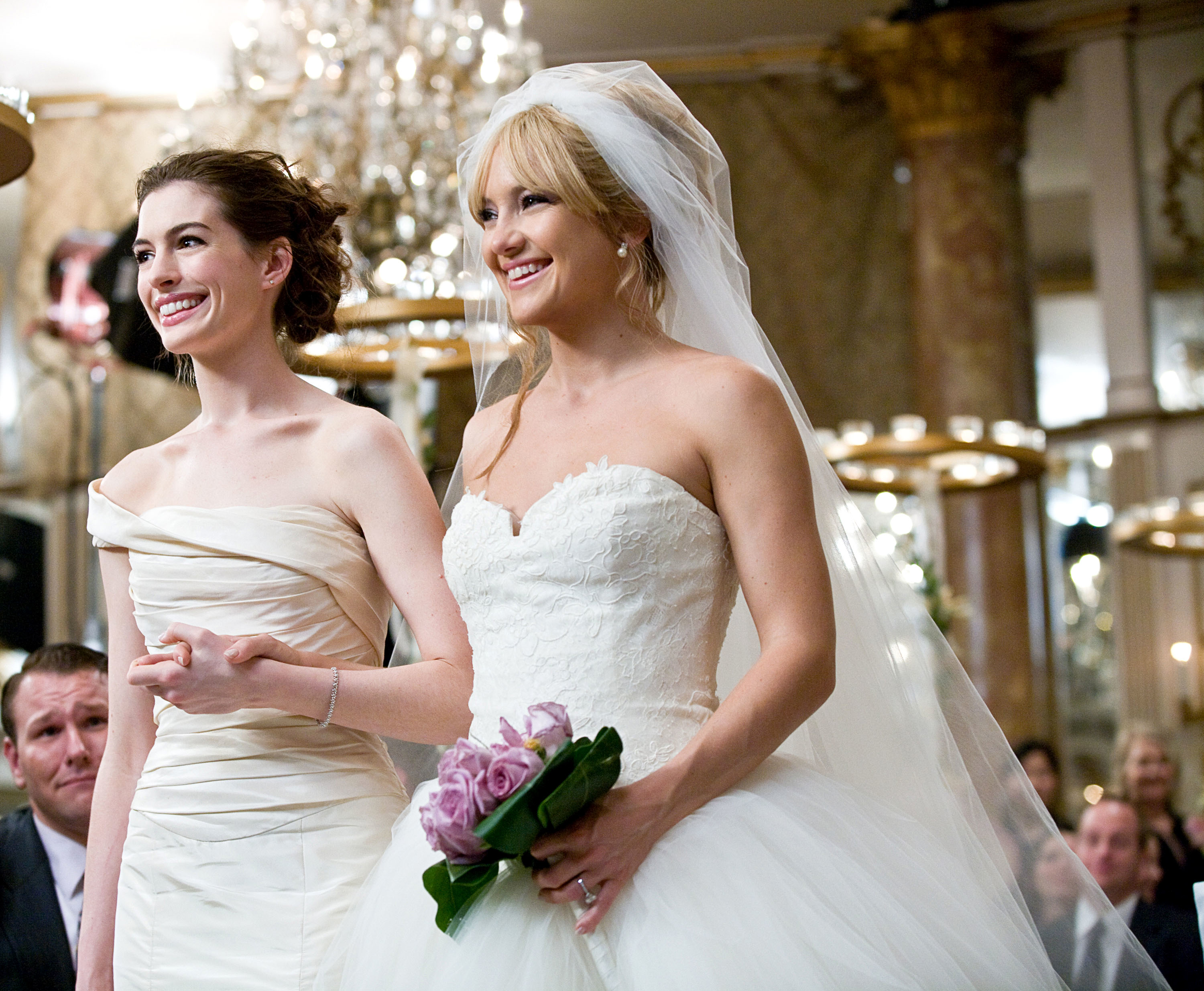 Anne Hathaway and Kate Hudson in wedding dresses, smiling and standing arm-in-arm at a wedding ceremony