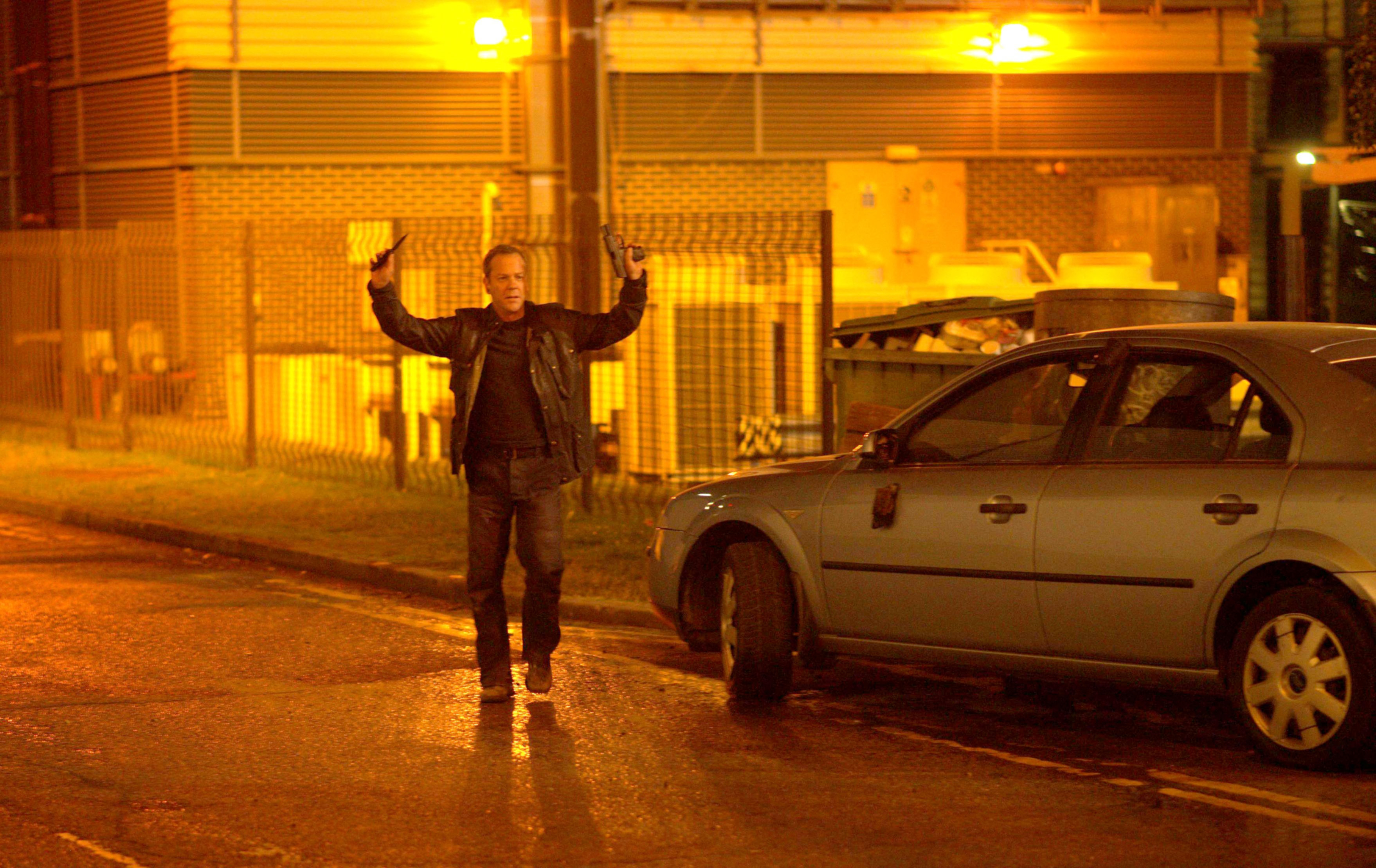 Kiefer Sutherland as Jack Bauer in a dramatic scene, holding two guns and standing beside a car at night