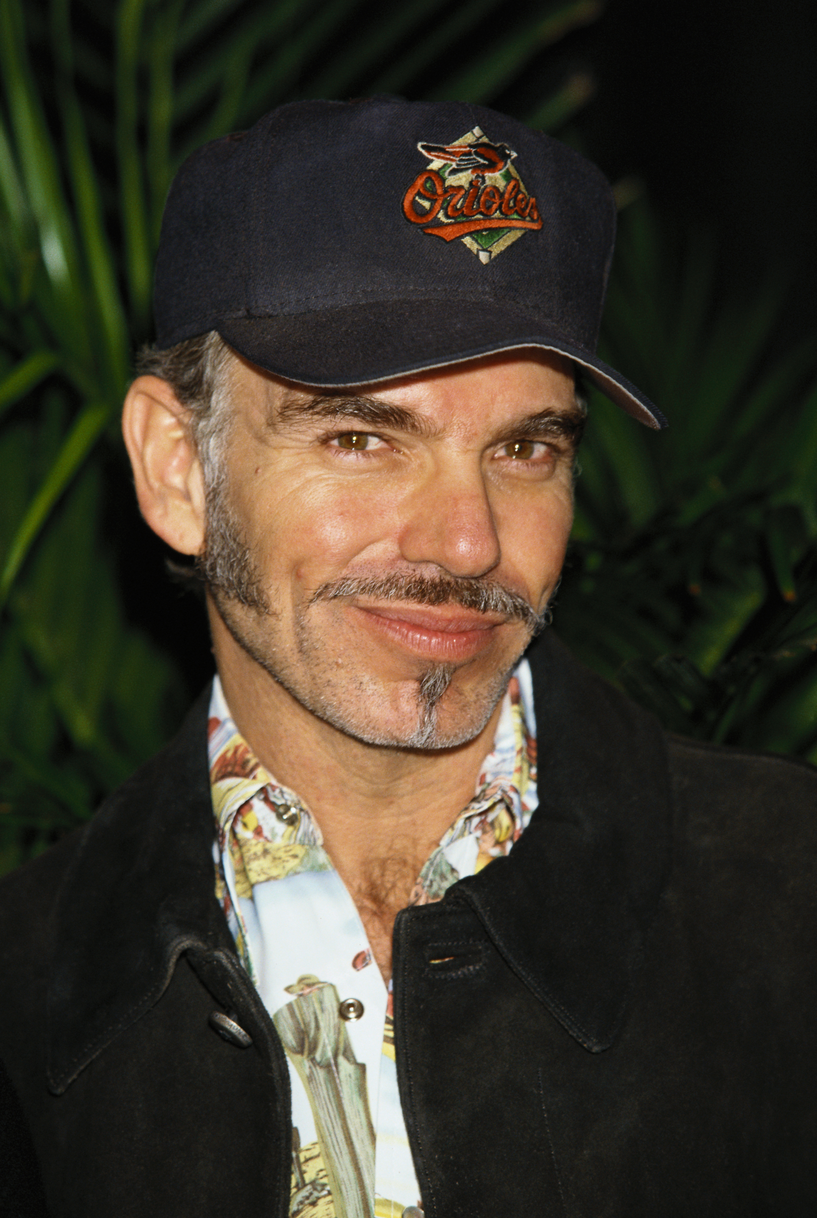 Billy Bob Thornton wears a baseball cap and a patterned shirt under a dark jacket, smiling at the camera