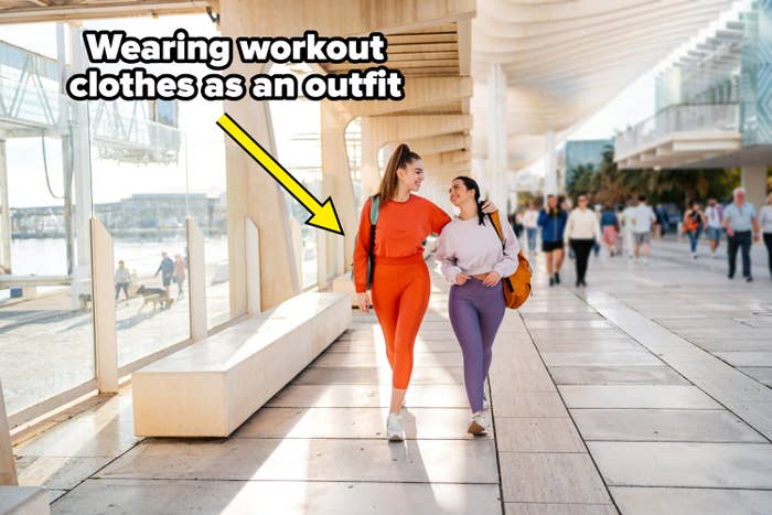 Two women wearing casual athletic wear walk side by side, smiling and chatting outdoors in a modern urban setting