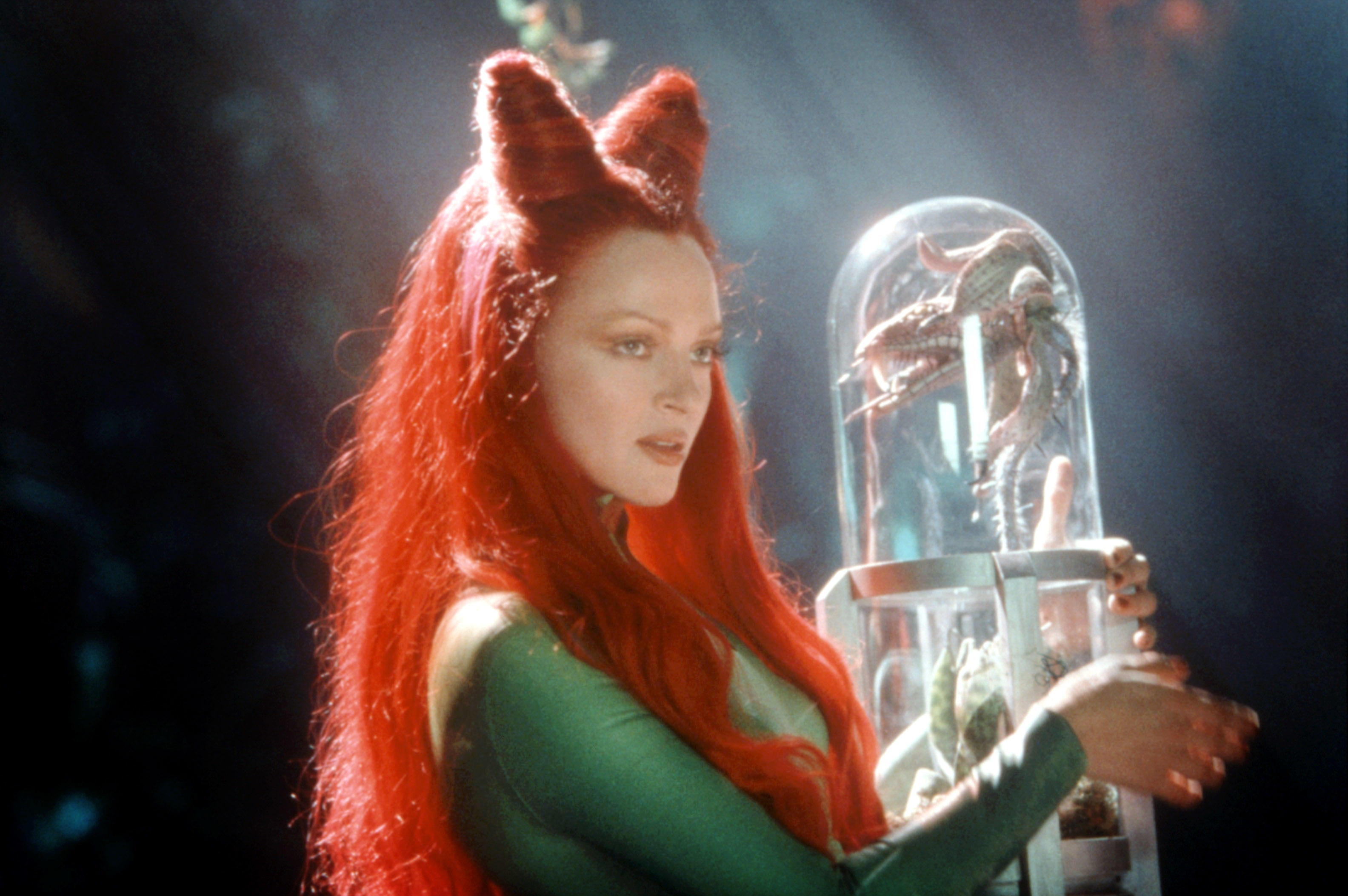 Uma Thurman dressed as Poison Ivy, in a green outfit with long red hair, holding a glass container with a plant inside in a scene from &quot;Batman &amp;amp; Robin.&quot;