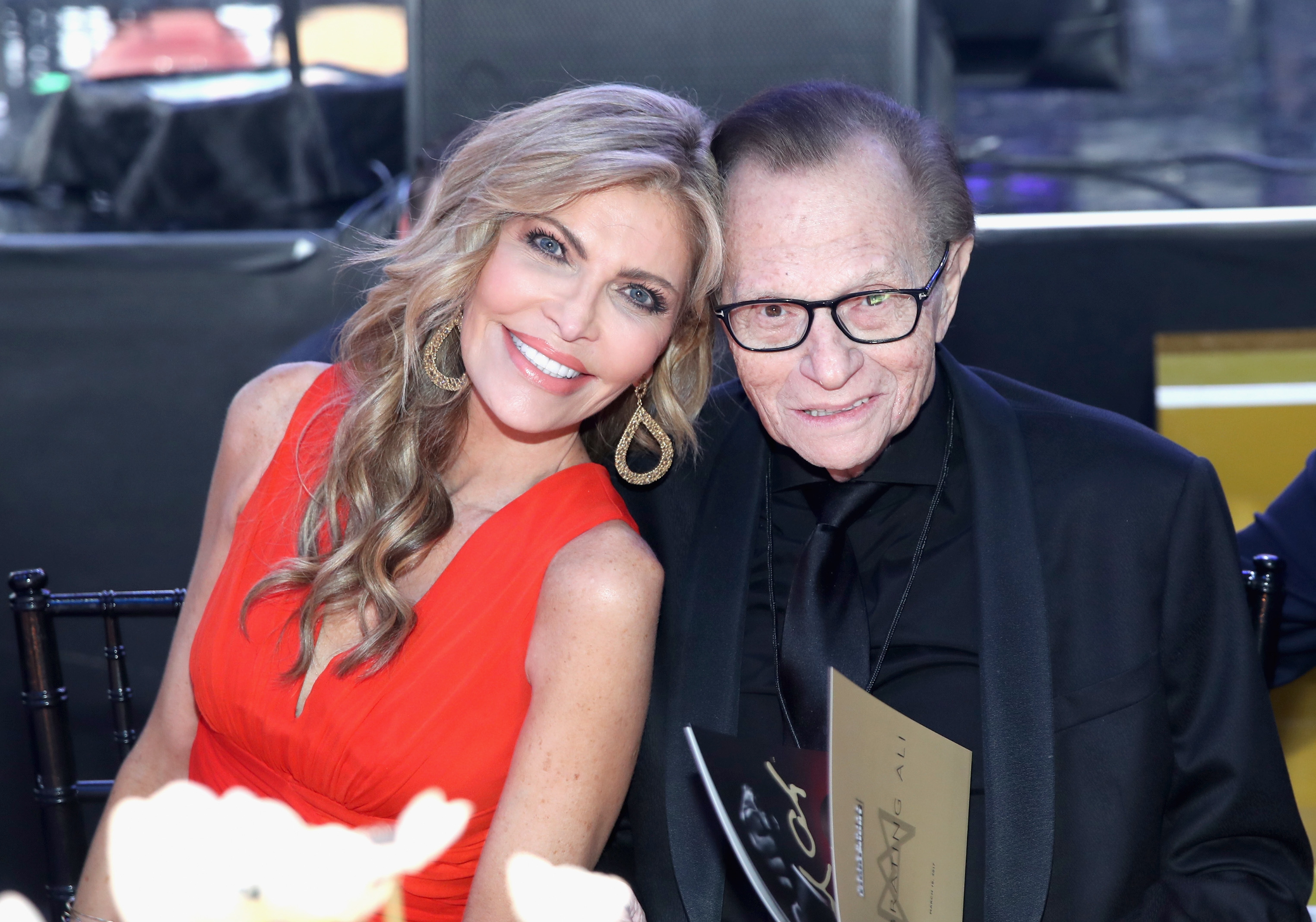 TV personality Larry King and his wife Shawn King sitting together at an event; Larry in a black suit and Shawn in an elegant sleeveless dress