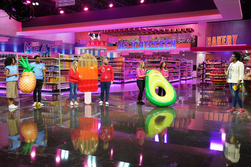 People on a game show stage that resembles a supermarket. Contestants hold large inflatable food items: ice cream, avocado, and popsicle. Host interacts with them