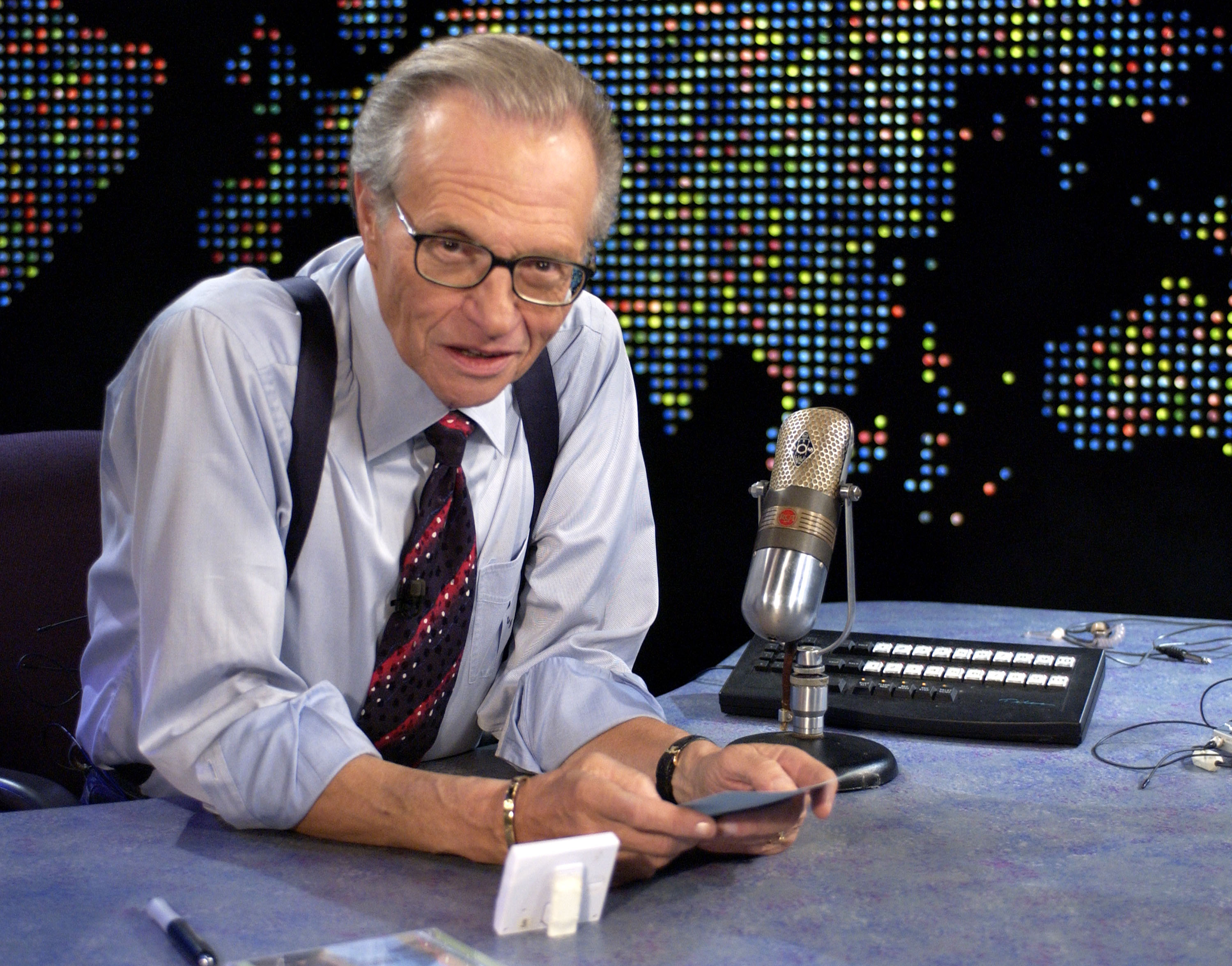 Larry King, wearing a shirt, tie, and suspenders, sits at a desk in a studio with a large microphone, ready for a broadcast