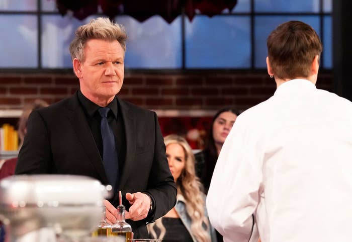 Gordon Ramsay in a dark suit and tie stands on a kitchen set, engaging with a person in a white chef's jacket. Other individuals are blurred in the background