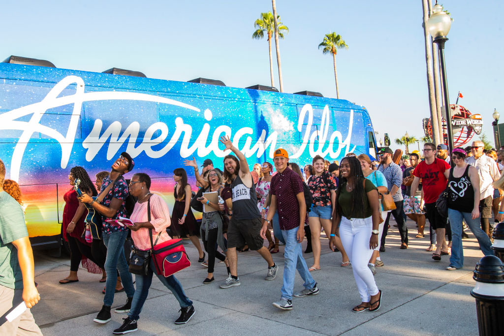 Group of people walking near a bus with &quot;American Idol&quot; on its side. They look excited and are smiling, wishing to participate in the show