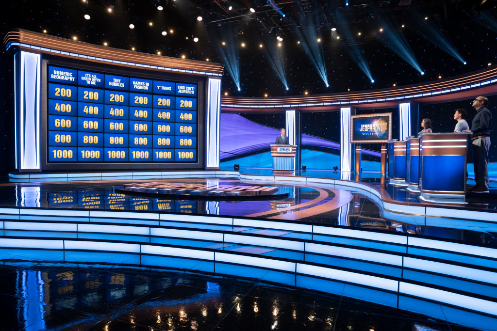 A Jeopardy game show set showcasing a board with categories and values, the host at a podium, and three contestants ready to answer questions